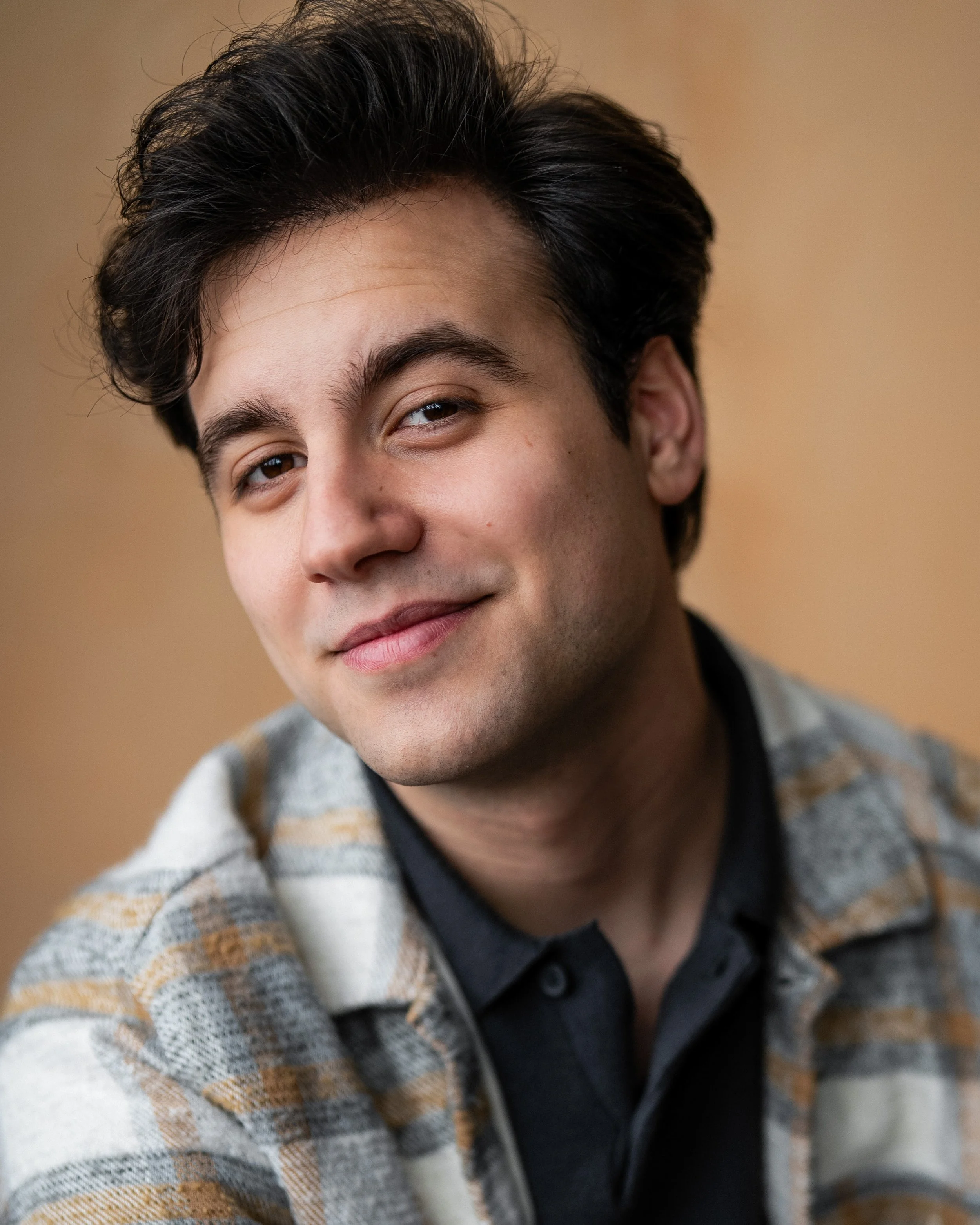 Matt Woodward, a young actor with dark brown hair and dark brown eyes, in a navy polo shirt with a tan and gray flannel over it, against a tan backdrop
