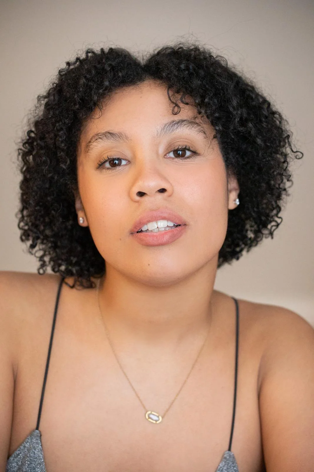 Close-up of Sawyer Evans, a young woman with curly dark hair, brown eyes, wearing a spaghetti strap top, earrings, and a gold necklace with a small oval pendant.