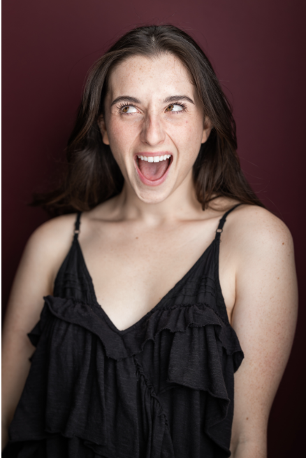 A woman with brown hair and freckles, wearing a black spaghetti strap top, expressing surprise or excitement against a dark red background.