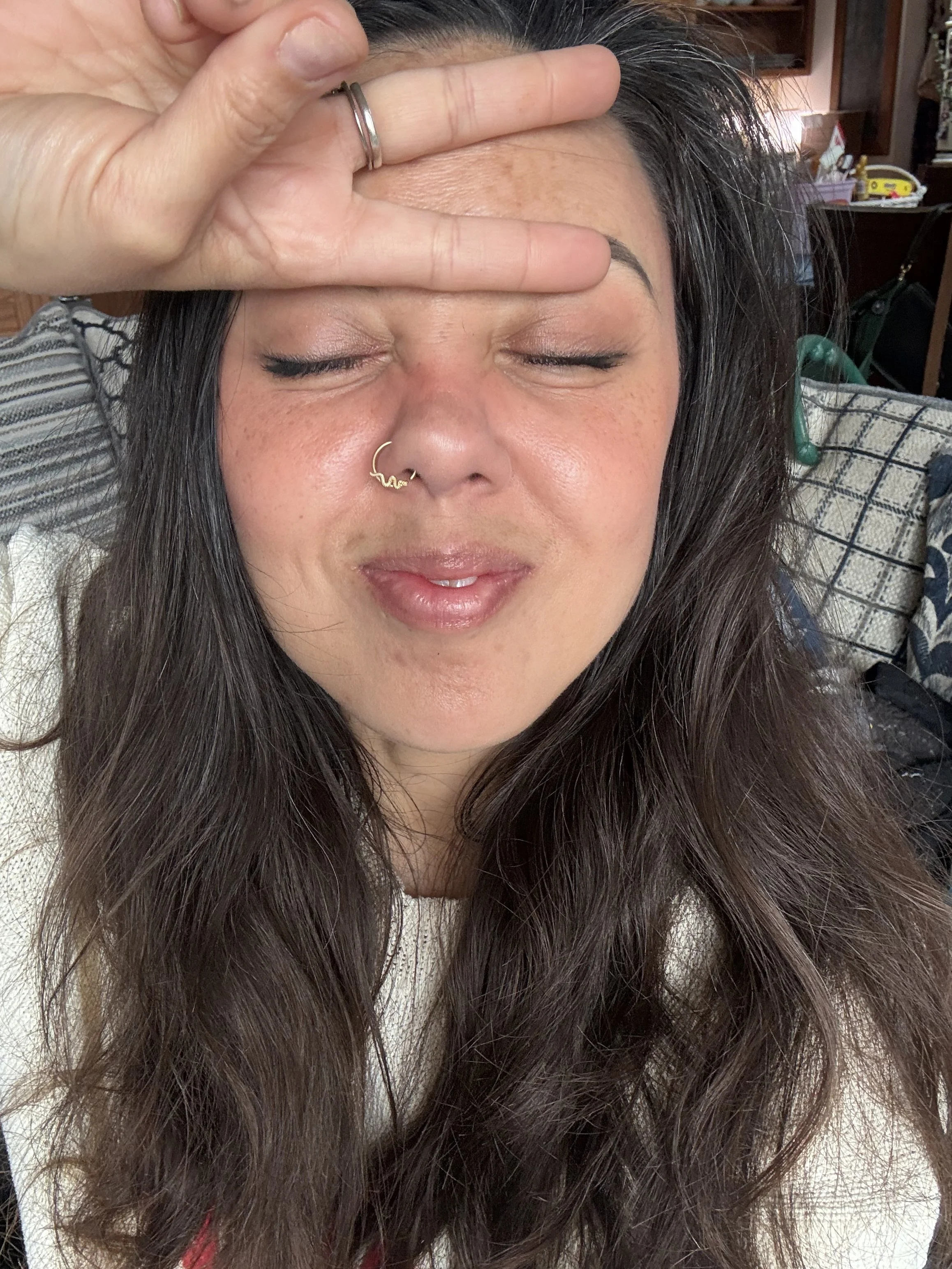 A woman with long dark hair, closed eyes, and a nose ring, resting her hand on her forehead.