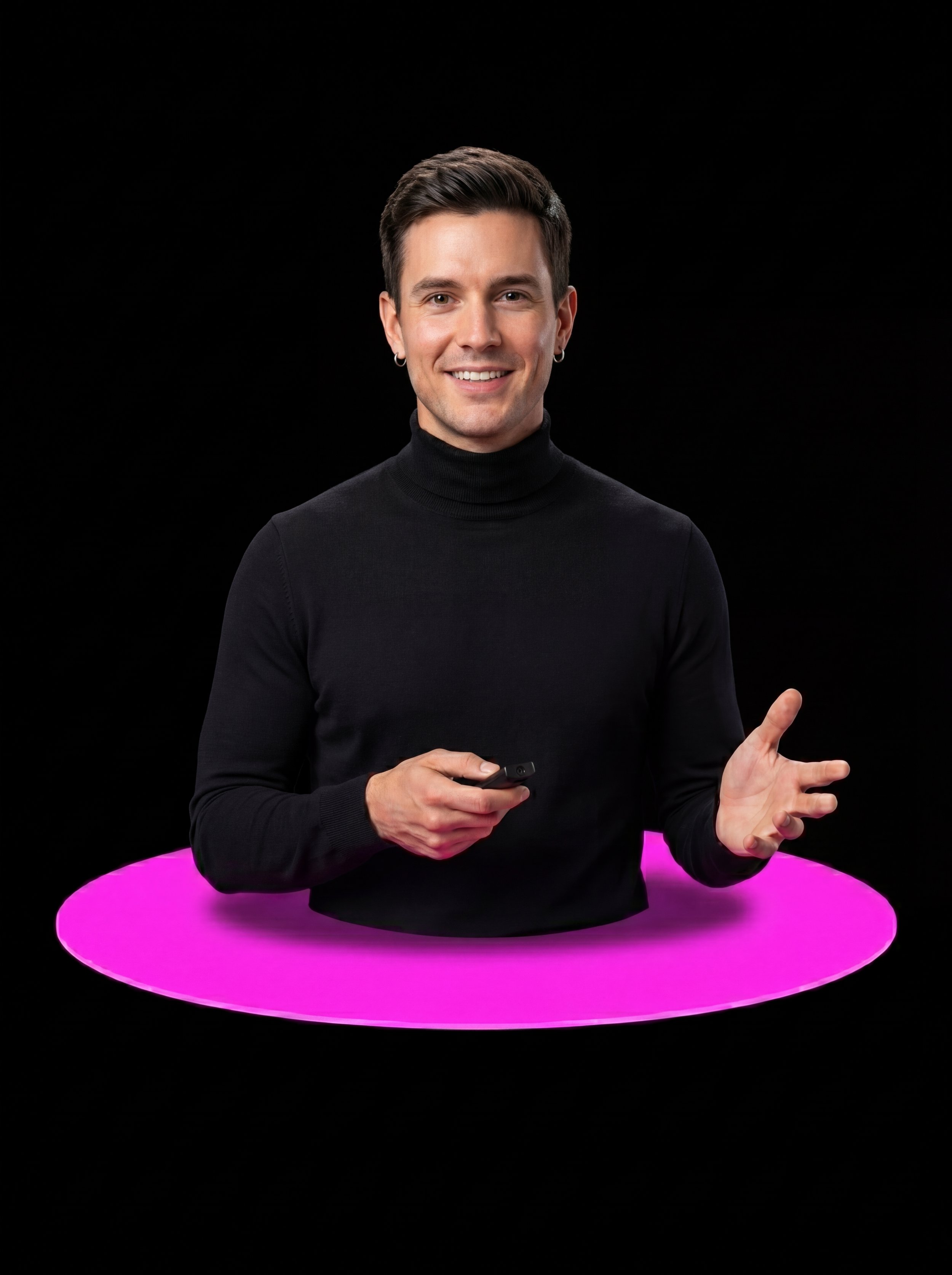 A young man with short dark hair, wearing a black turtleneck, smiling, holding a remote control, and gesturing with his left hand, standing behind a pink circular light on a black background.