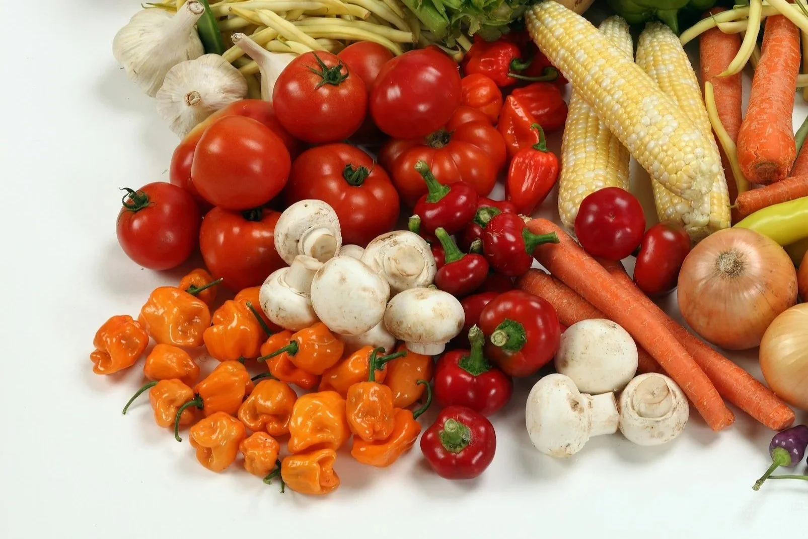 A variety of fresh vegetables including tomatoes, garlic, mushrooms, carrots, yellow corn, yellow peppers, and habanero peppers on a white surface.