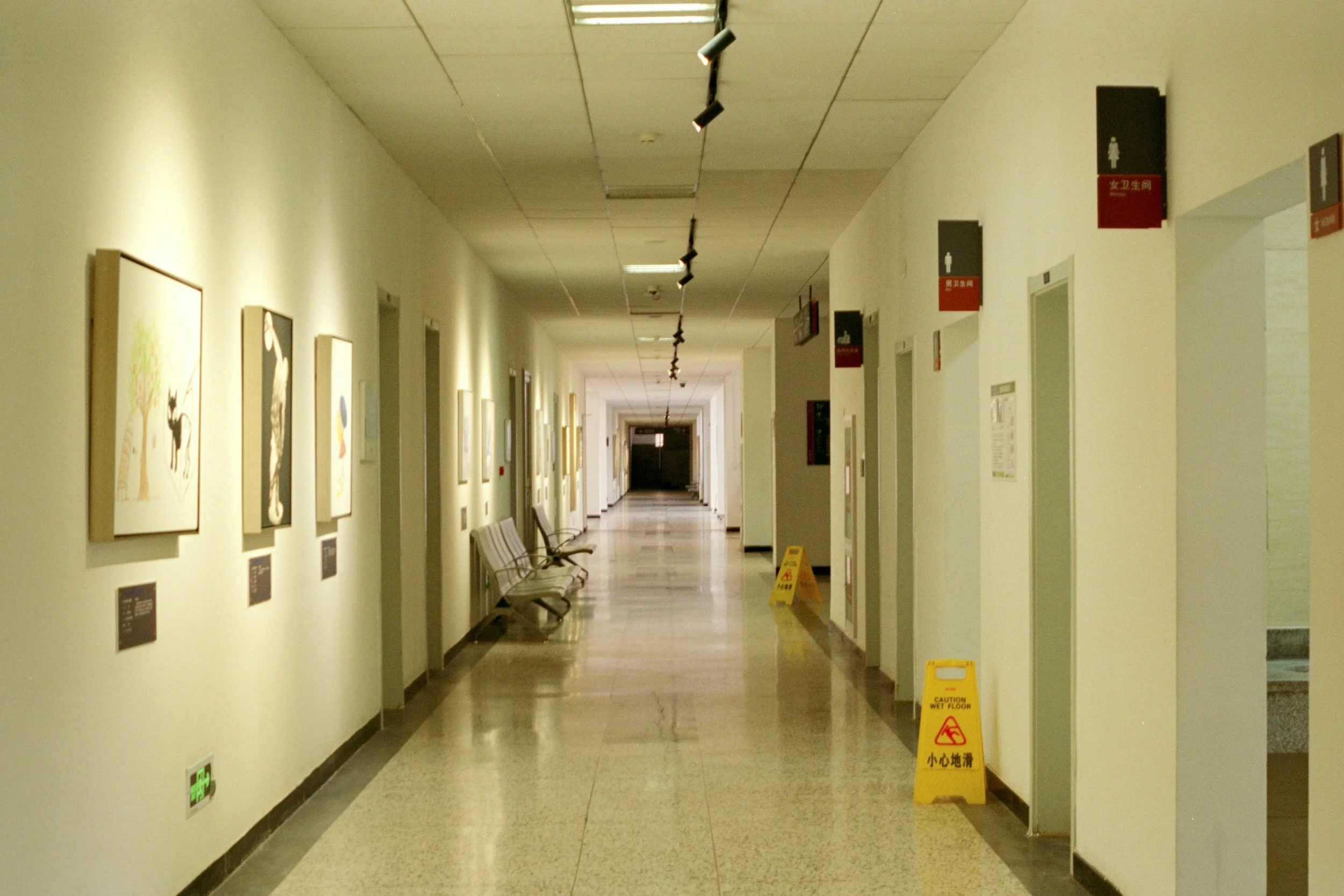 A long, empty hallway in a building with framed artwork on the left wall and advertisement signs on the right wall. Several chairs are positioned along the left side, and yellow caution signs indicating a wet floor are placed on the floor. The ceiling has black track lighting and fluorescent lights.