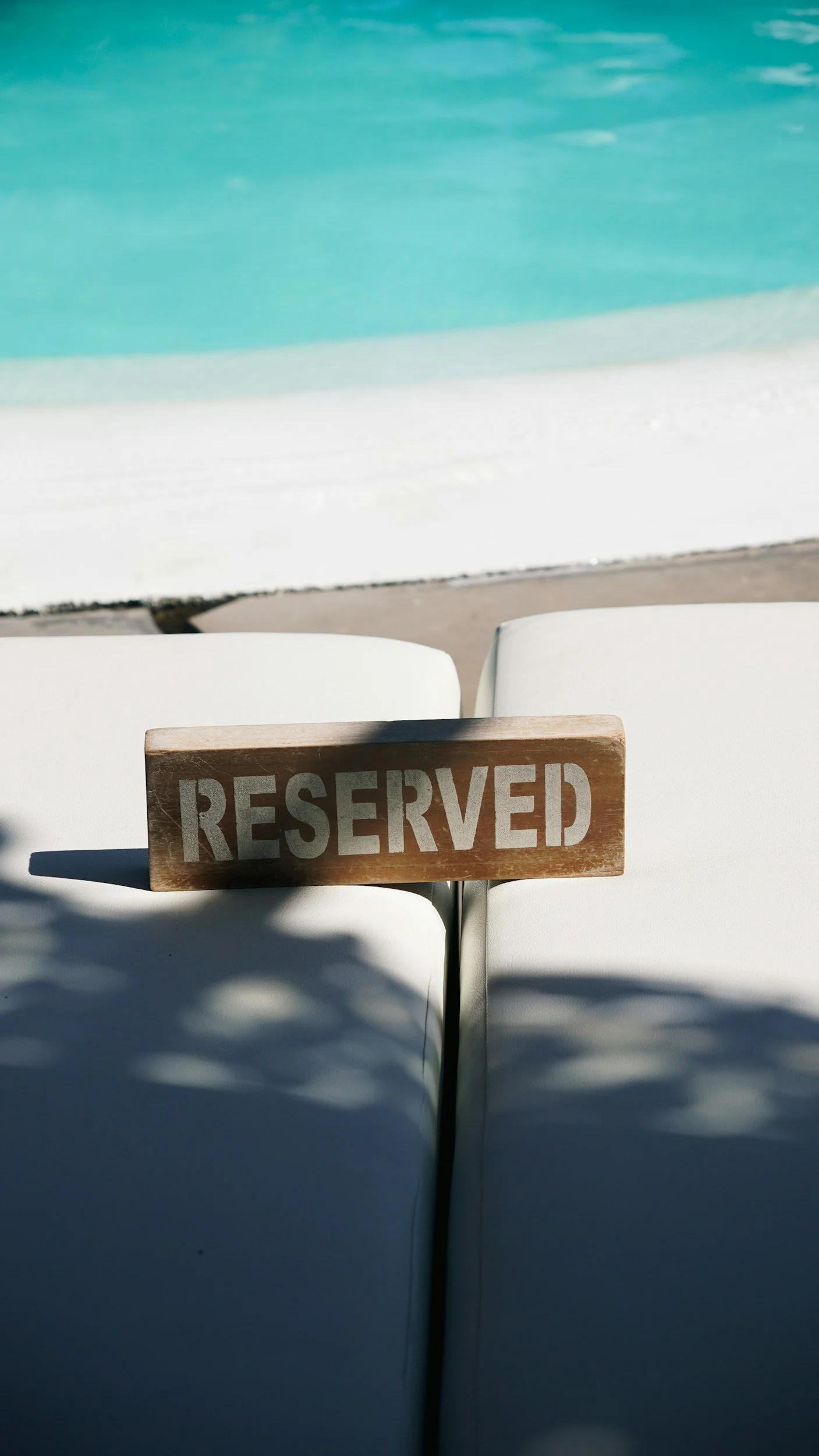 Two white beach chairs with a wooden sign that reads 'RESERVED' placed on one of them, near a swimming pool with clear turquoise water.