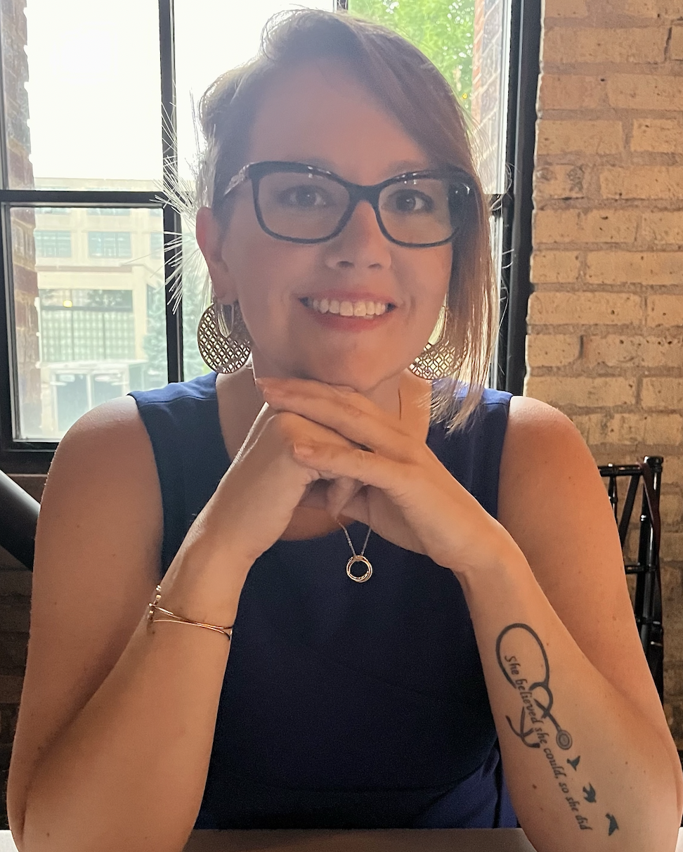 A woman sitting at a table in a café or restaurant with brick walls and large windows, smiling and resting her chin on her clasped hands. She has short brown hair, glasses, and is wearing a navy blue sleeveless top, circular earrings, and a pendant necklace. She has a tattoo on her left forearm with text and bird illustrations.
