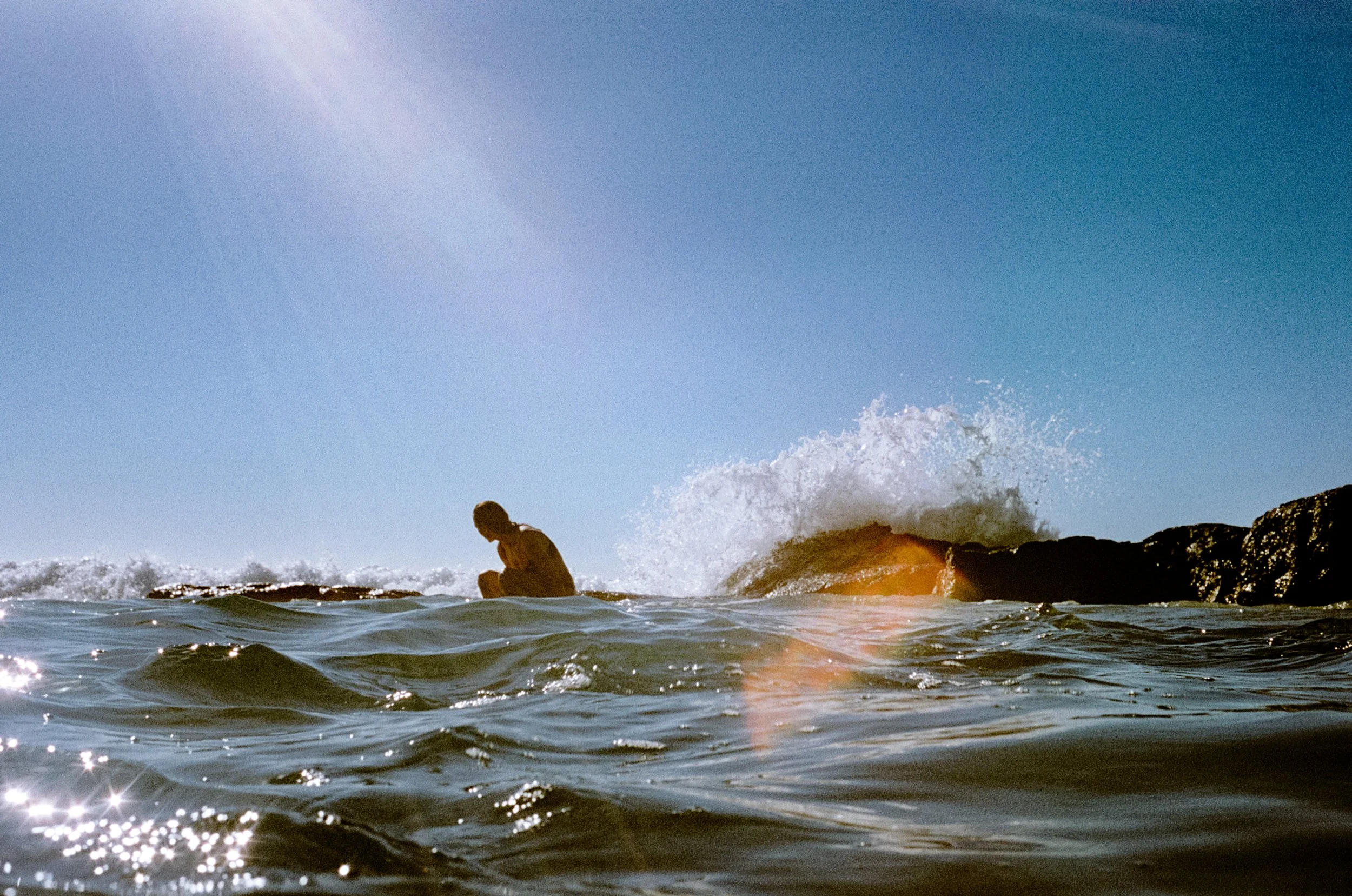 Lauren crouched on coastal rocks as surf breaks around her, sun flare across the ocean, NSW South Coast.