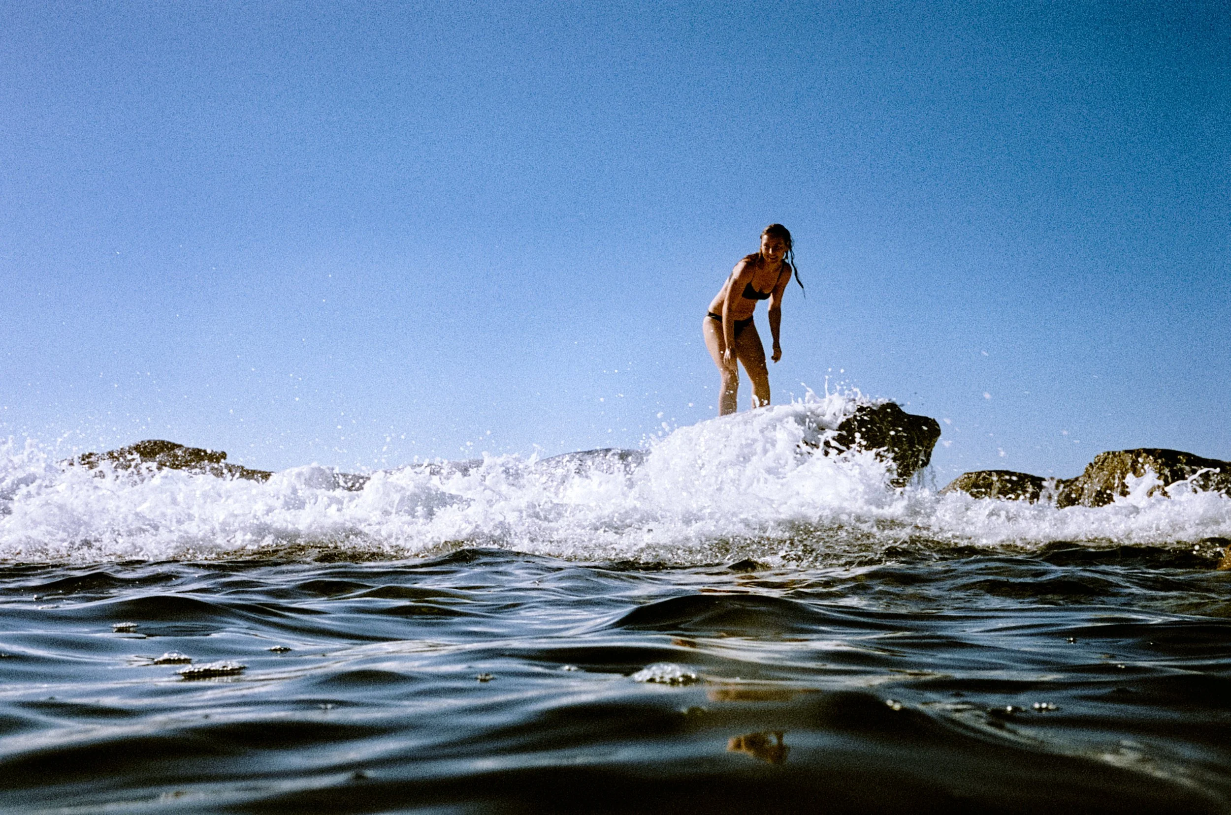 Lauren standing in ocean surf, waves washing around her legs, bright blue sky and rocky outcrops behind, NSW South Coast.