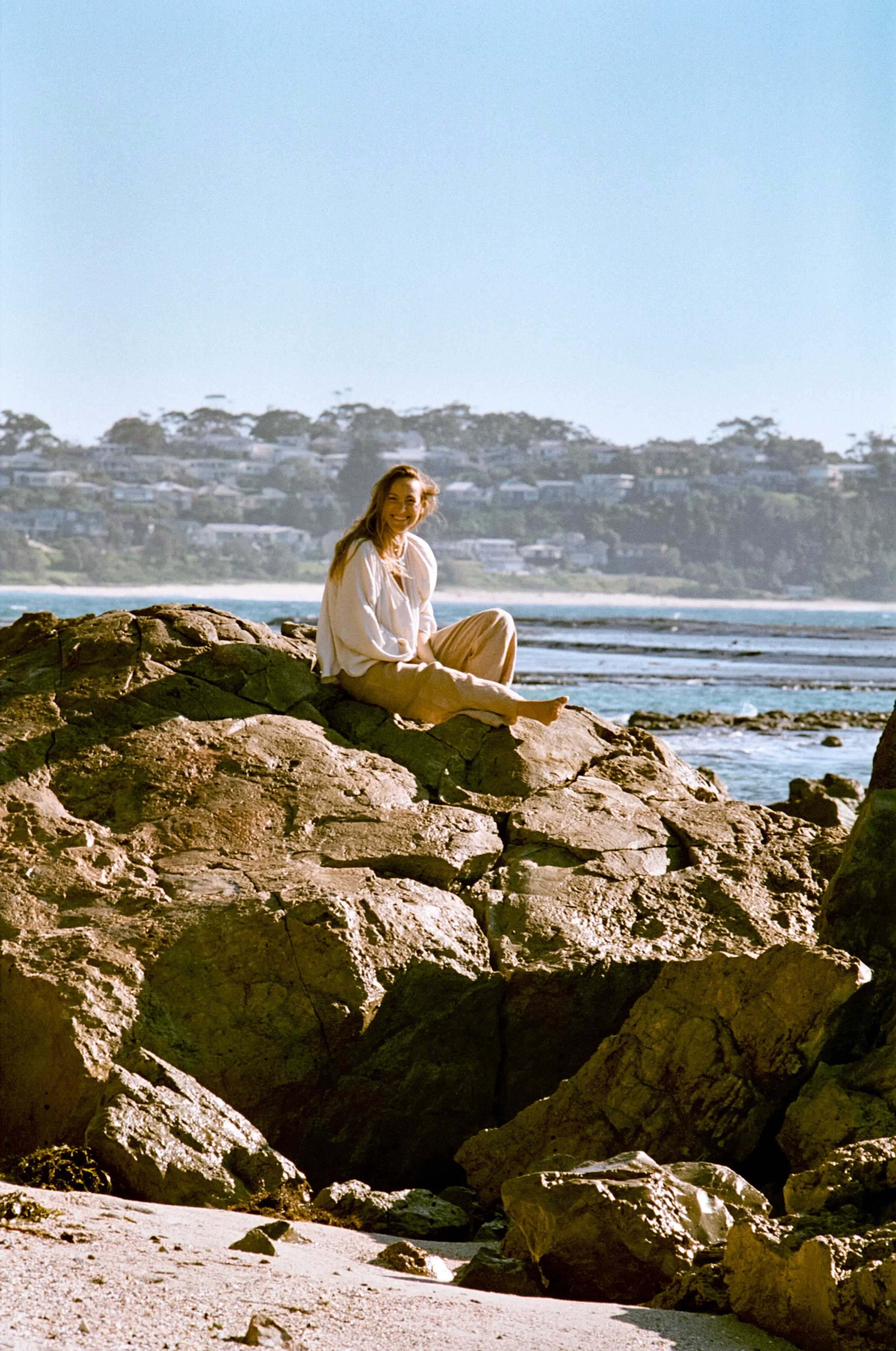 Lauren sitting on coastal rocks at the ocean's edge, headland and beach houses behind, NSW South Coast.