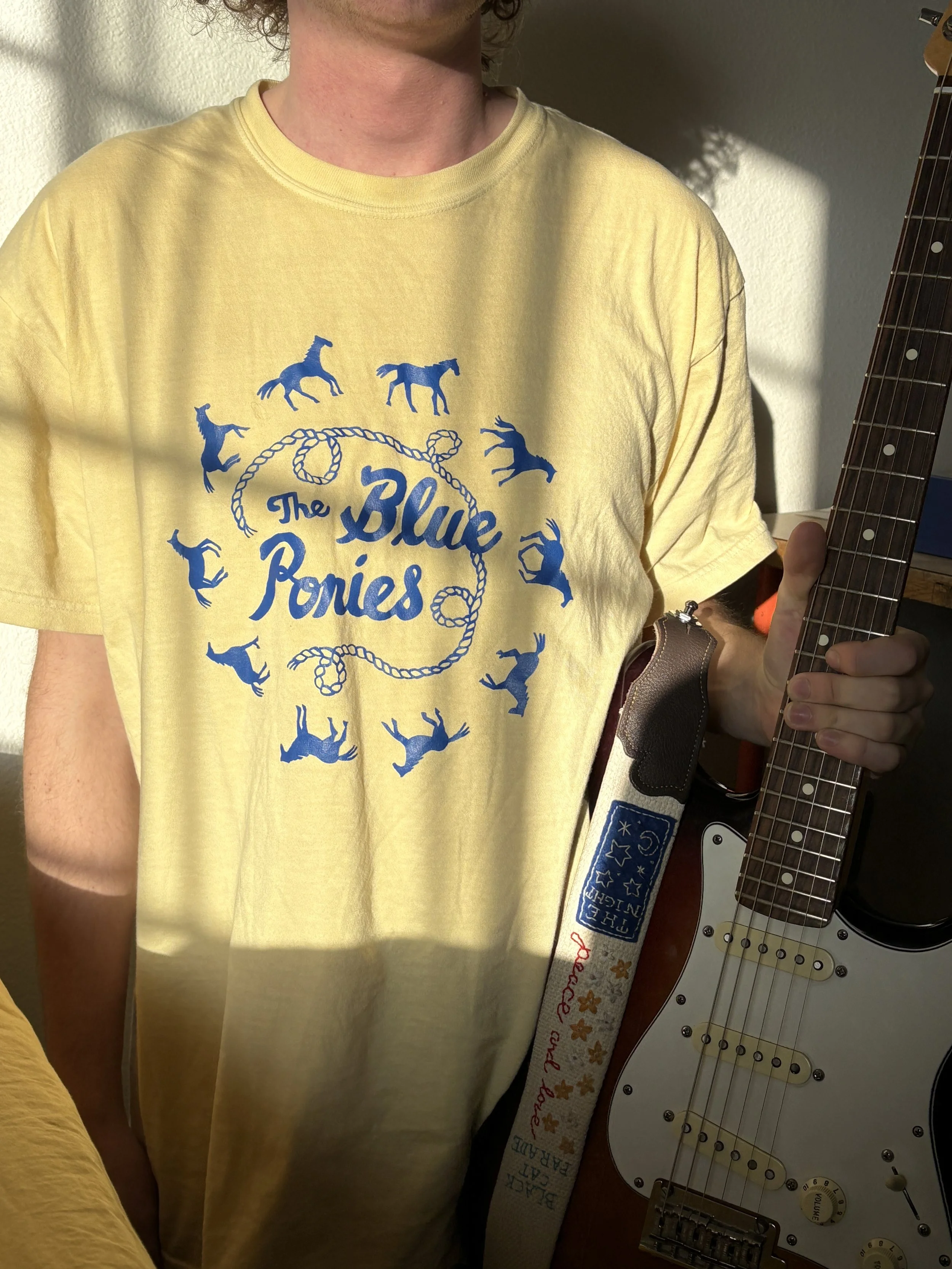 Person wearing a yellow T-shirt with blue text and graphics, holding an electric guitar.