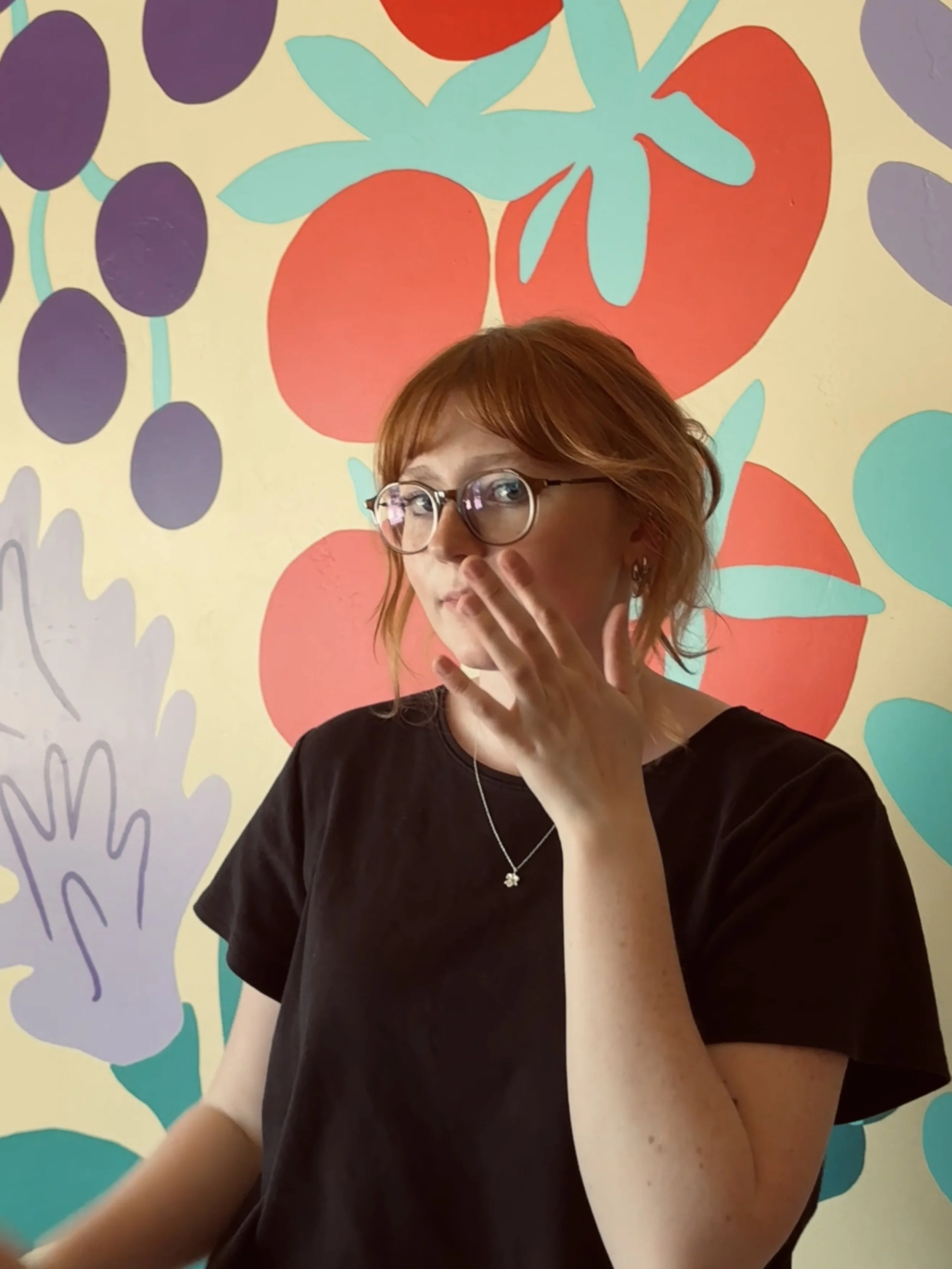 A woman with short red hair and glasses poses against a colorful, fruit-themed mural, with her hand near her face and a thoughtful expression.