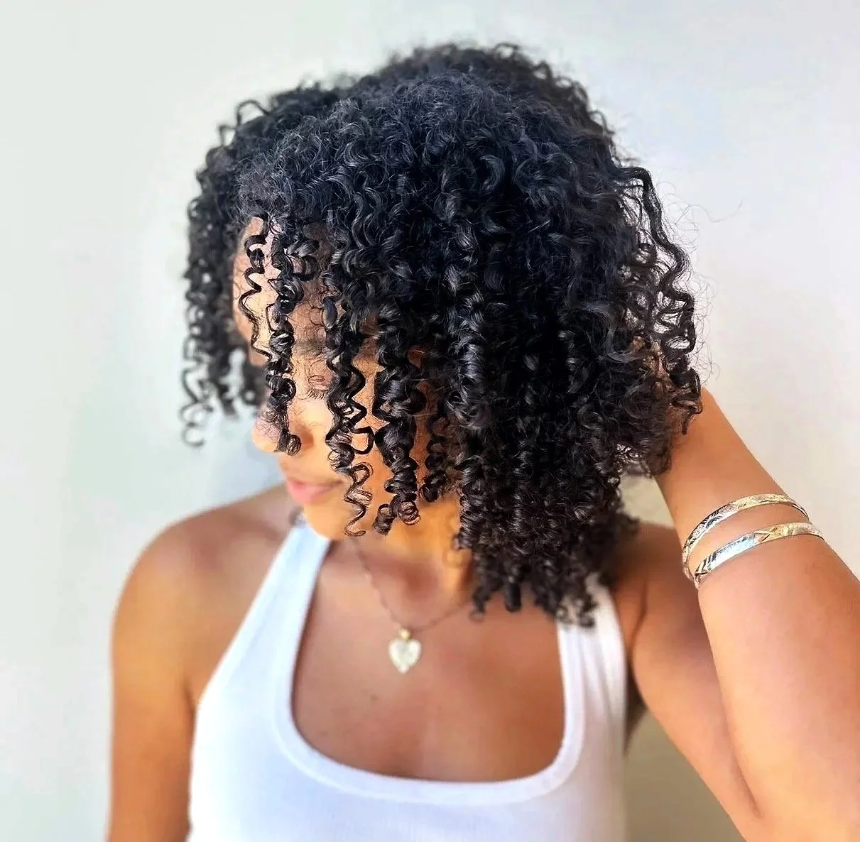 A woman with dark, curly hair styled in defined ringlets, wearing a white tank top, gold bracelets, and a heart-shaped necklace, with her hand touching her hair.