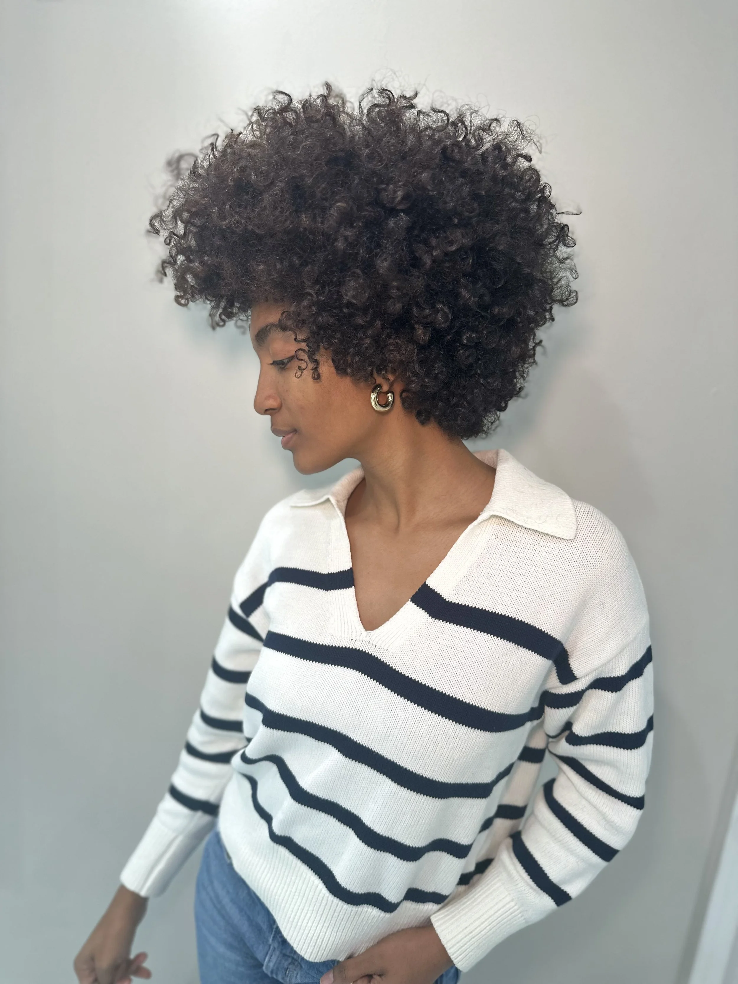Side view of a woman with curly dark hair wearing a white sweater with navy stripes and blue jeans, standing against a light gray wall.