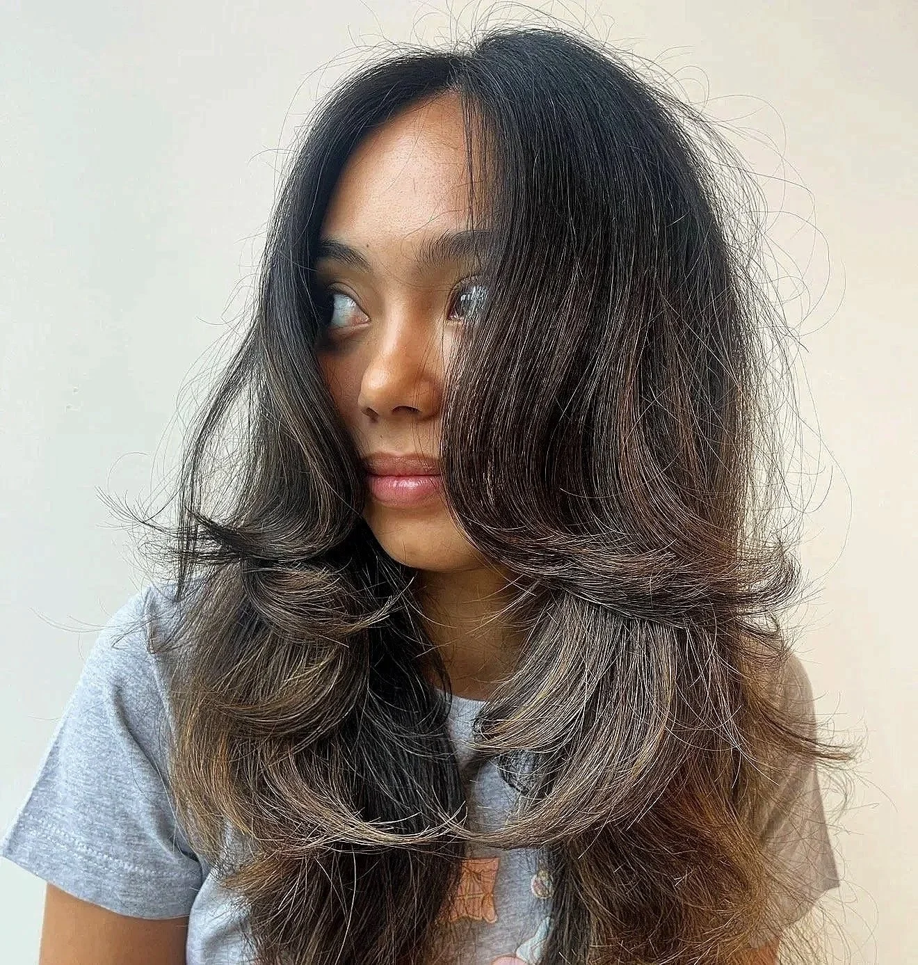 A woman with long, wavy dark hair with silver highlights, looking to her left against a plain light-colored background.