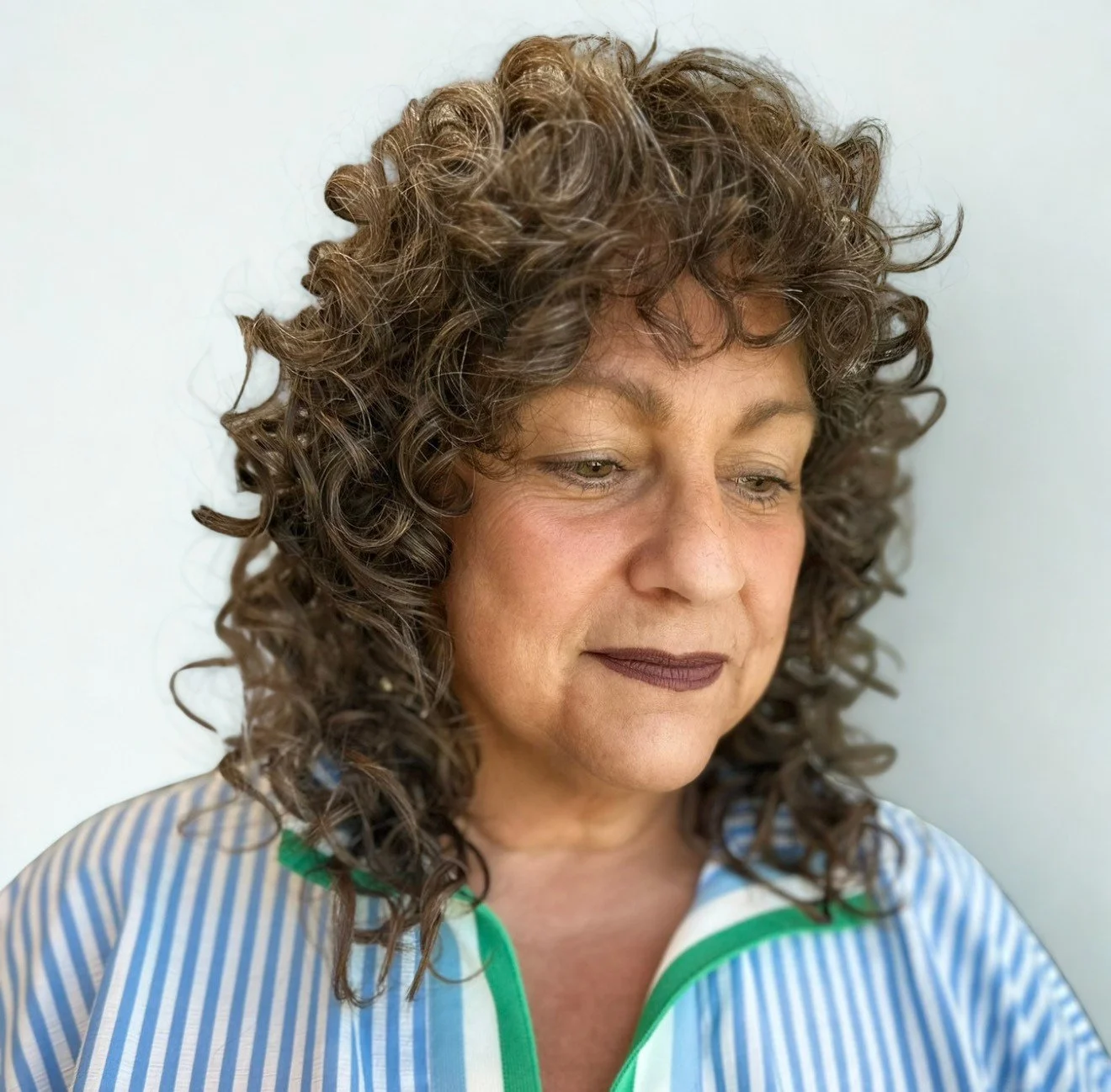 A woman with curly brown hair, wearing a blue and white striped shirt with green accents, looking down with a neutral expression.