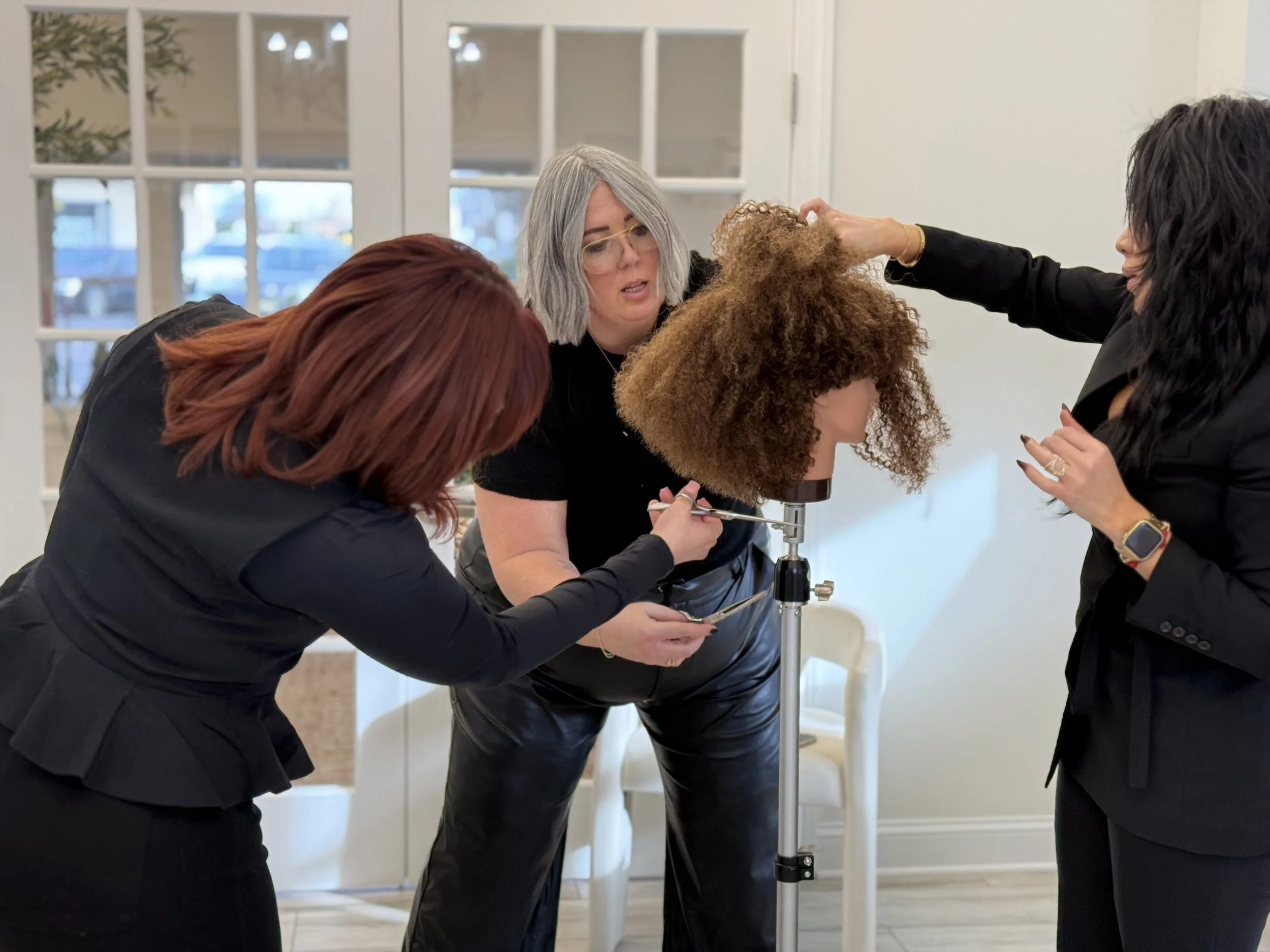 Four women with hair colors red, gray, curly and black are examining a wig on a stand in a bright, modern room.