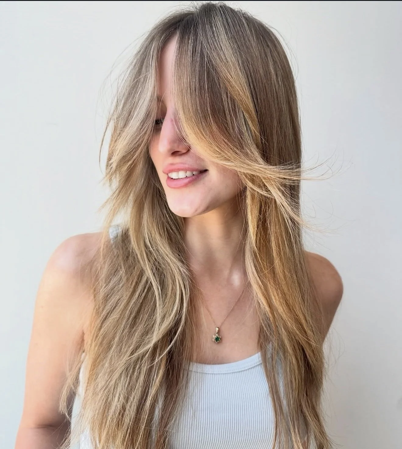 A woman with long, wavy blonde hair smiling, wearing a white tank top and a necklace with a green pendant.