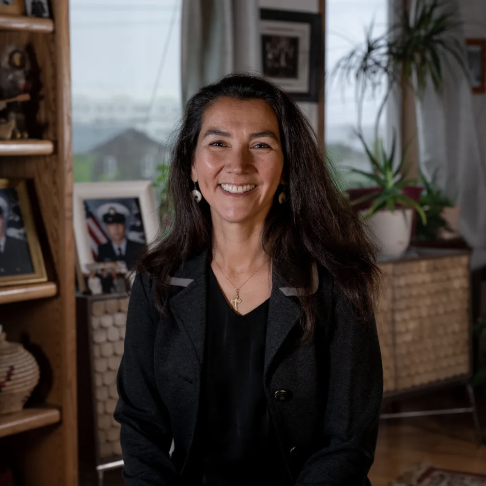 A smiling woman with long dark hair, wearing a black blazer, jewelry, and a gold cross necklace, sitting in a cozy room with framed photographs, plants, and wooden furniture.
