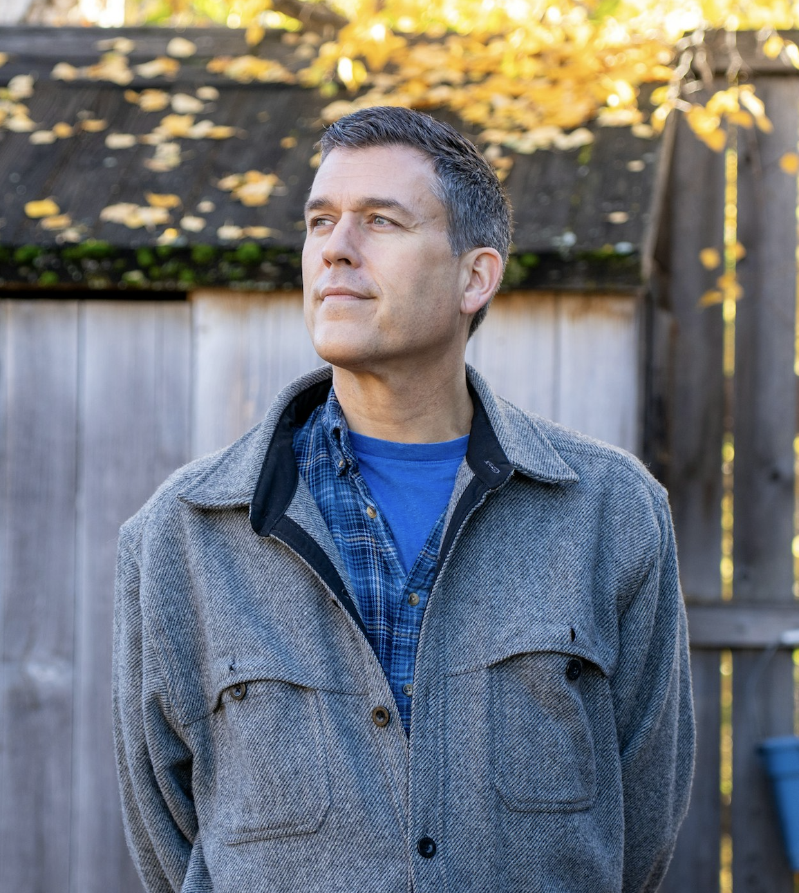 A man standing outdoors in front of a wooden fence with autumn leaves on the trees behind.