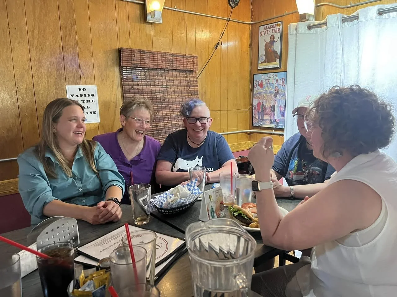 Six women seated around a restaurant table laughing and talking. There are drinks, food, and a basket with napkins on the table. The background features wood-paneled walls, a sign that says 'No vaping in this area,' and posters for Alaska State Fair. A light curtain covers a window.