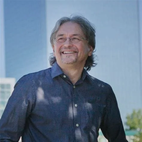 A smiling man with gray hair and glasses, wearing a dark blue button-up shirt, standing outdoors with a cityscape in the background.