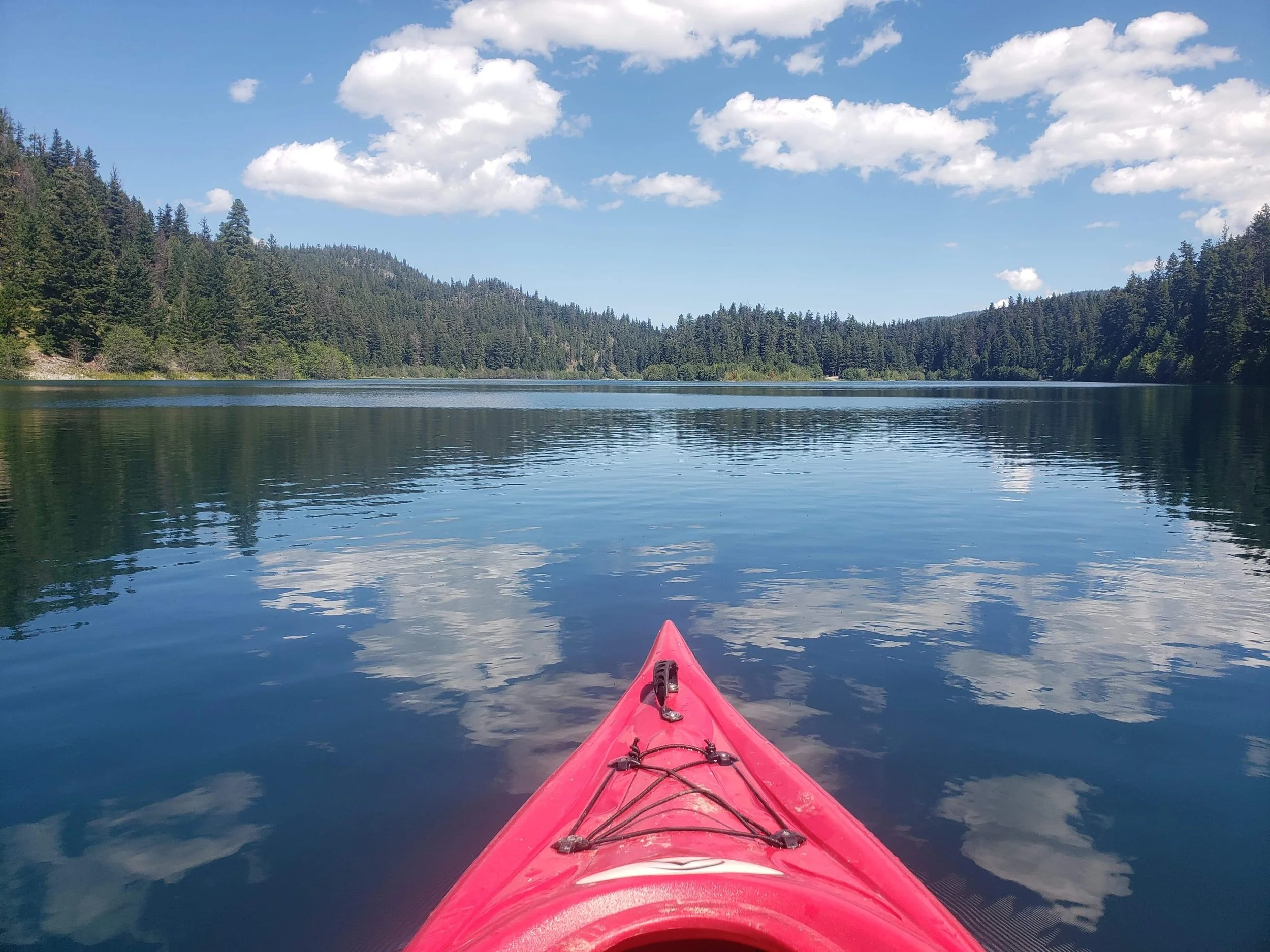 View from a kayak on a calm mountain lake, surrounded by lush green trees and forested hills, with a partly cloudy sky above.