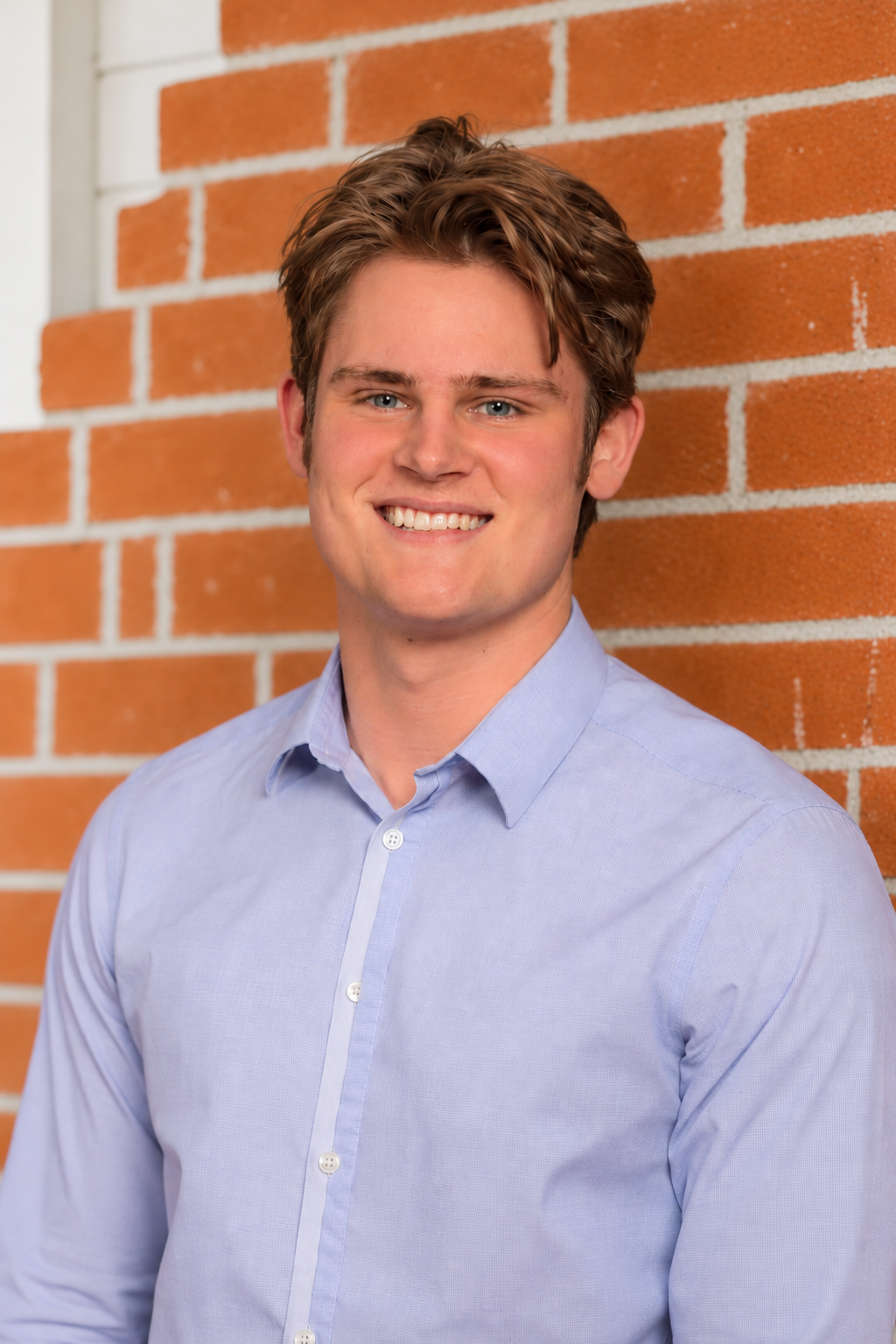 Smiling young man in a light blue button-down shirt standing against a red brick wall
