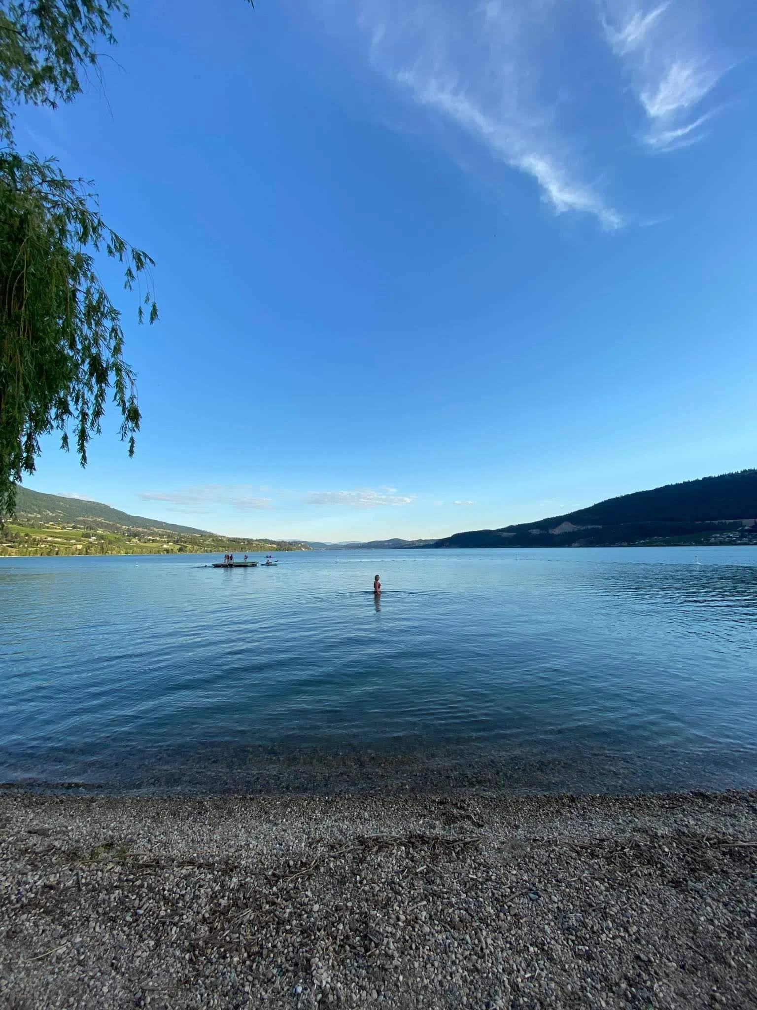 A view of a large lake with a pebble beach in the foreground, green hills in the background, a blue sky with some clouds, a boy standing in the water, and a boat with people on it.