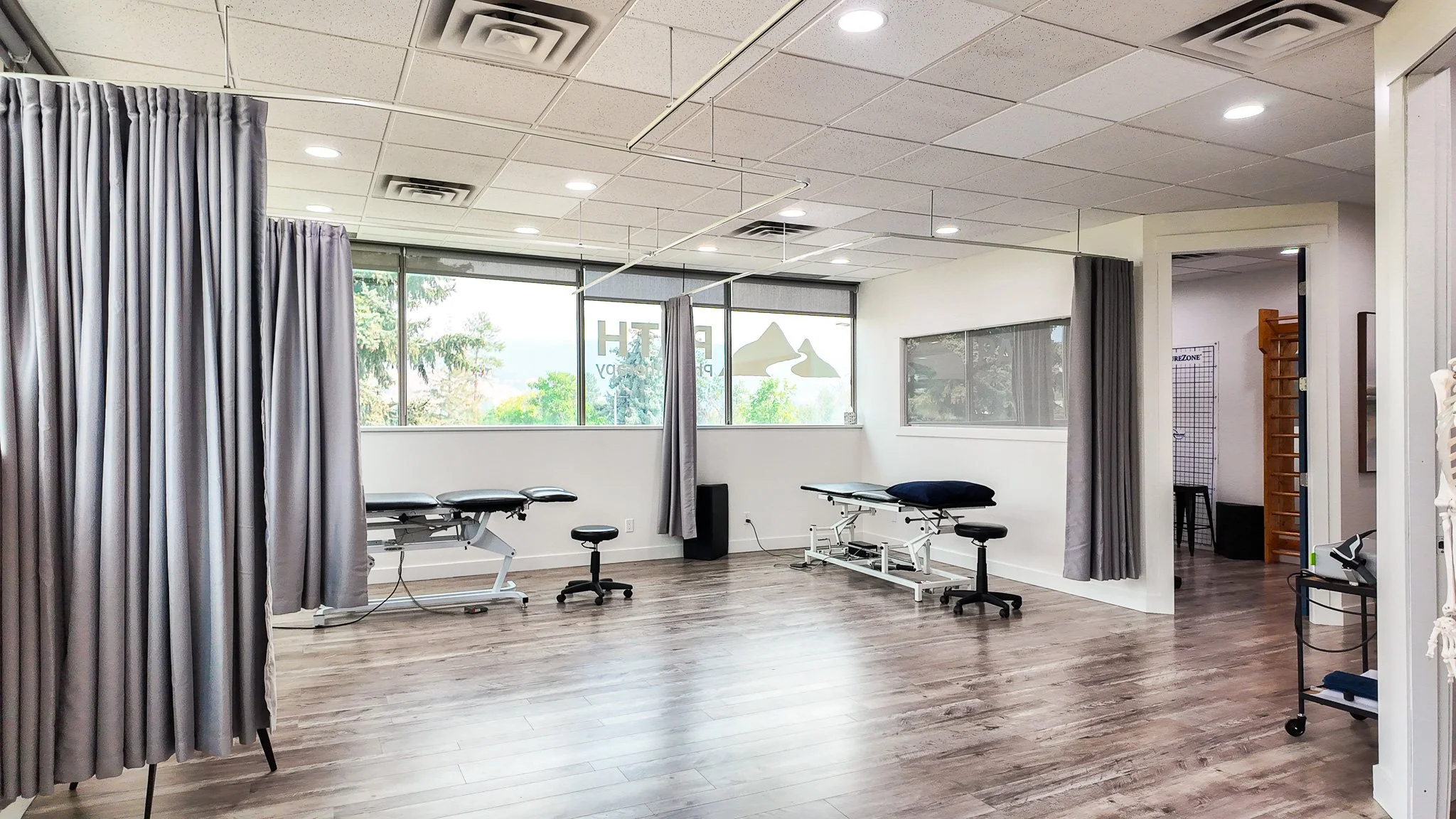Bright physical therapy clinic with treatment tables, stools, privacy curtains, and exercise equipment.