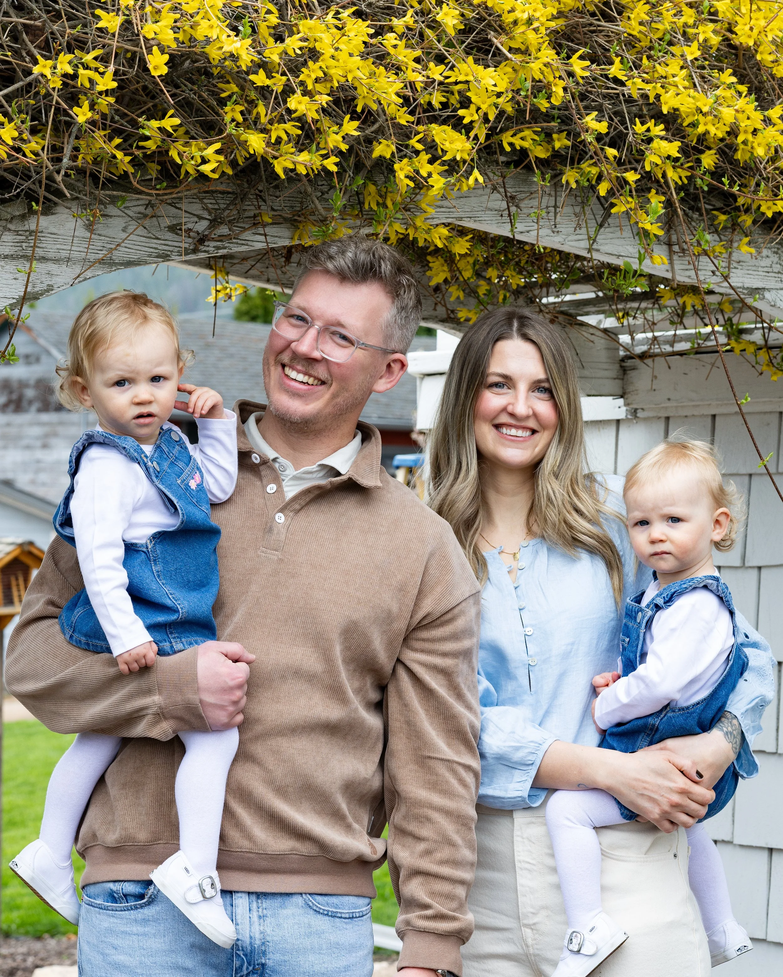A smiling family of four outdoors, with two parents holding their young daughters, standing under a yellow flowering vine on a trellis.