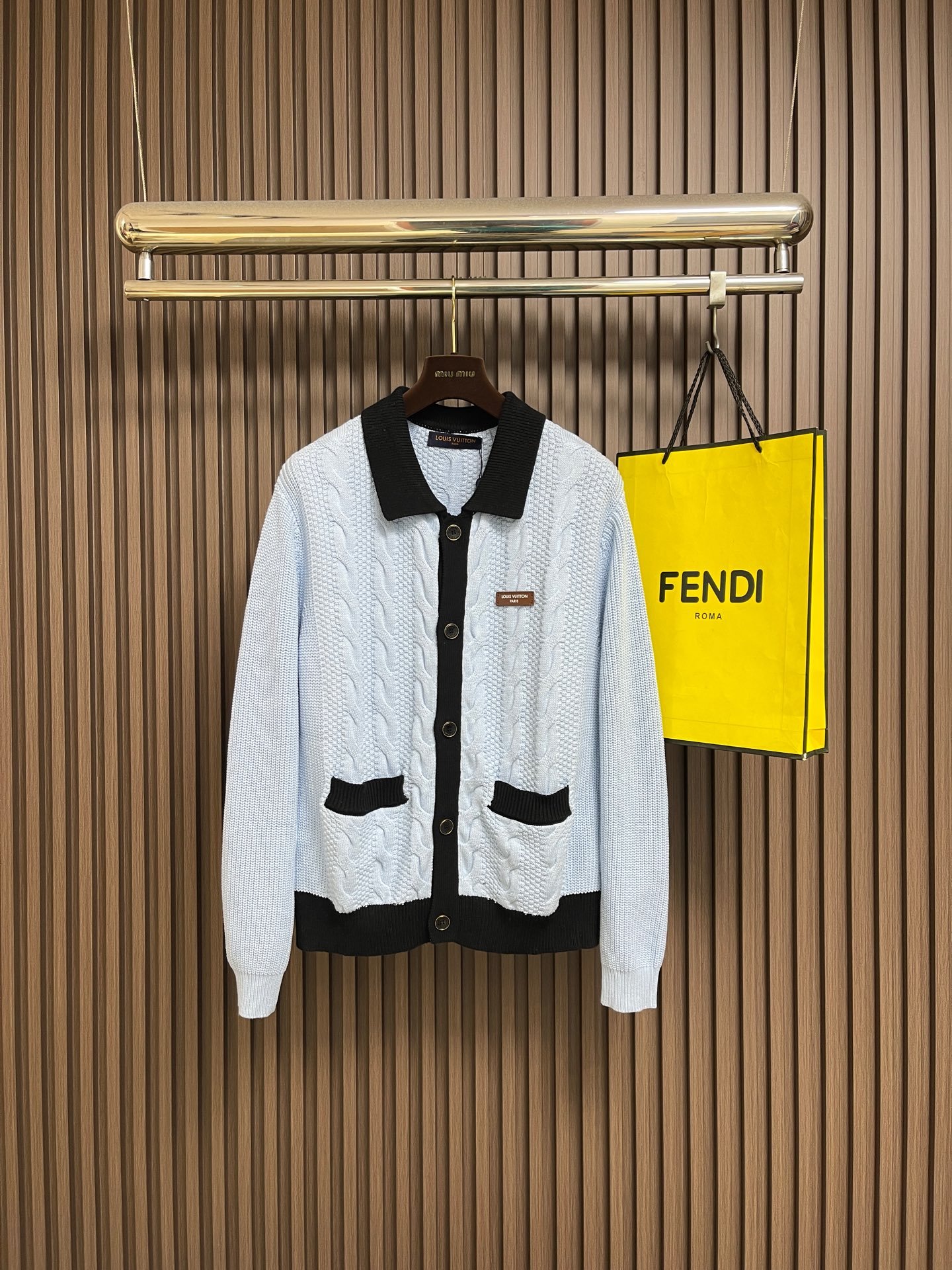 A white knitted cardigan with black trim on the collar, pockets, and buttons hanging on a black hanger, with a yellow Fendi shopping bag beside it, set against a wooden slat wall background.