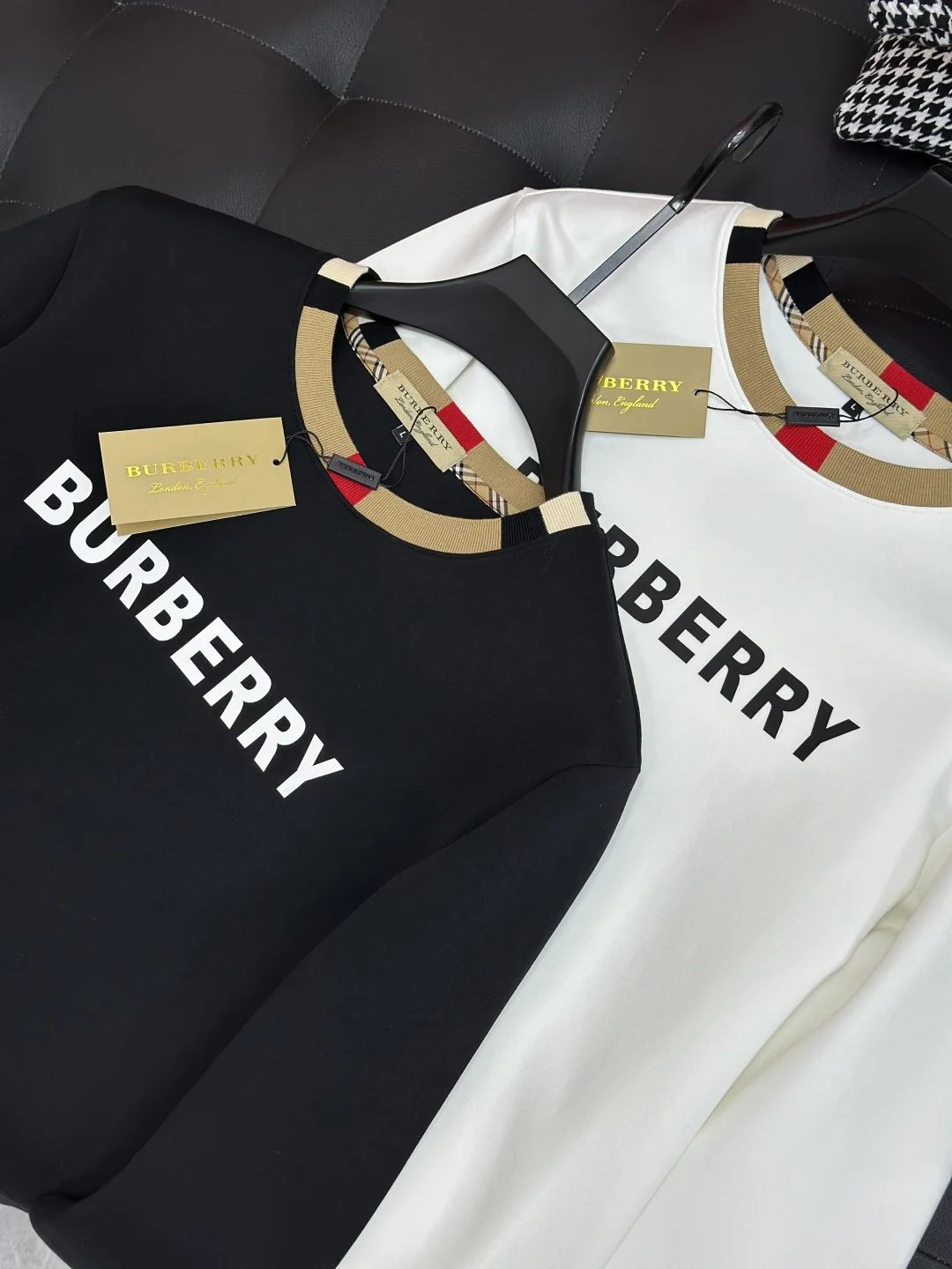 Two Burberry sweatshirts, one black and one white, with the Burberry logo printed on the front, hanging on black hangers with tags, on a dark background.