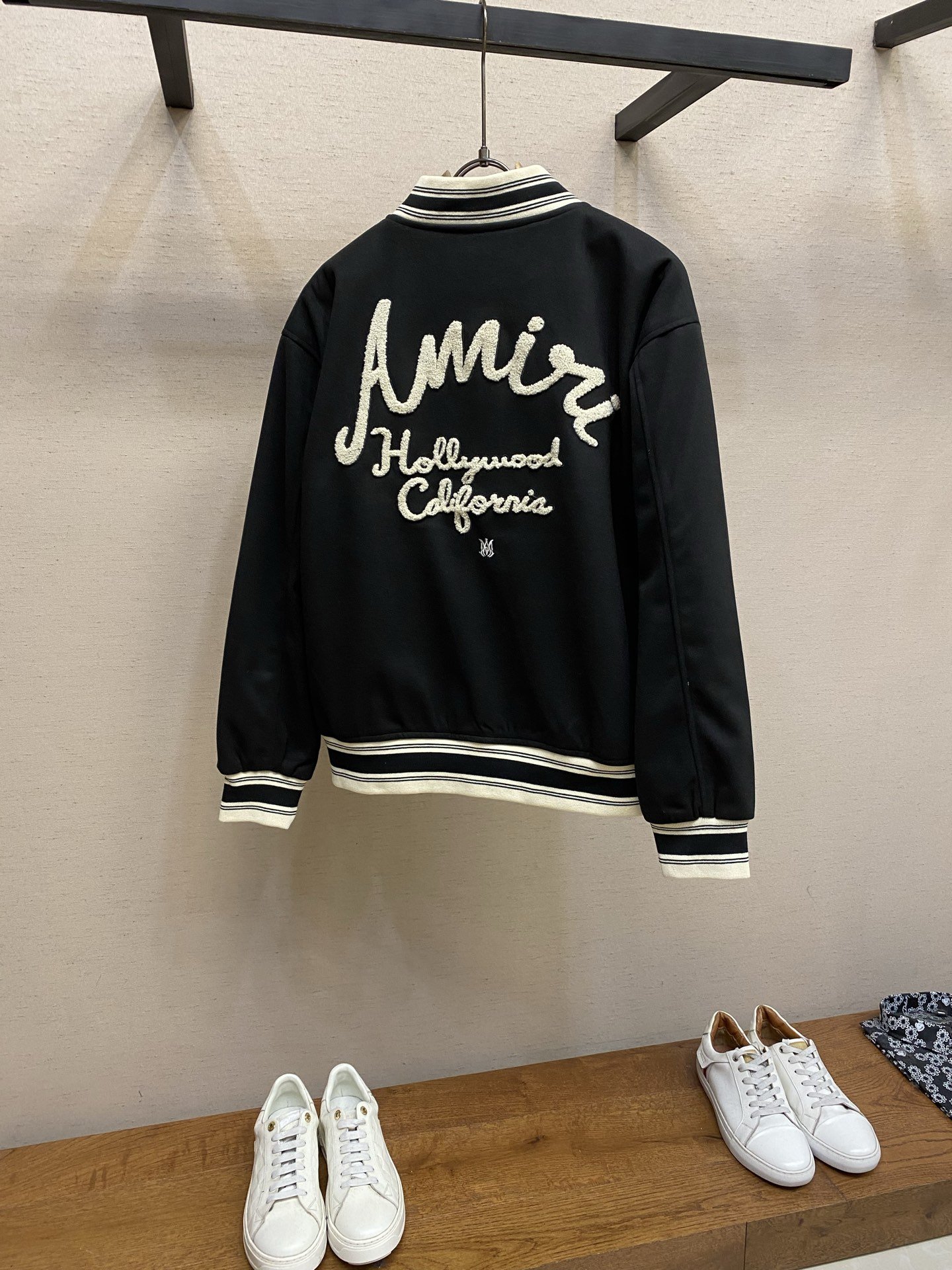 Black jacket with white embroidered text on the back that says 'Amizi Hollywood California,' hanging on a hanger, with pairs of white sneakers on a wooden table below.
