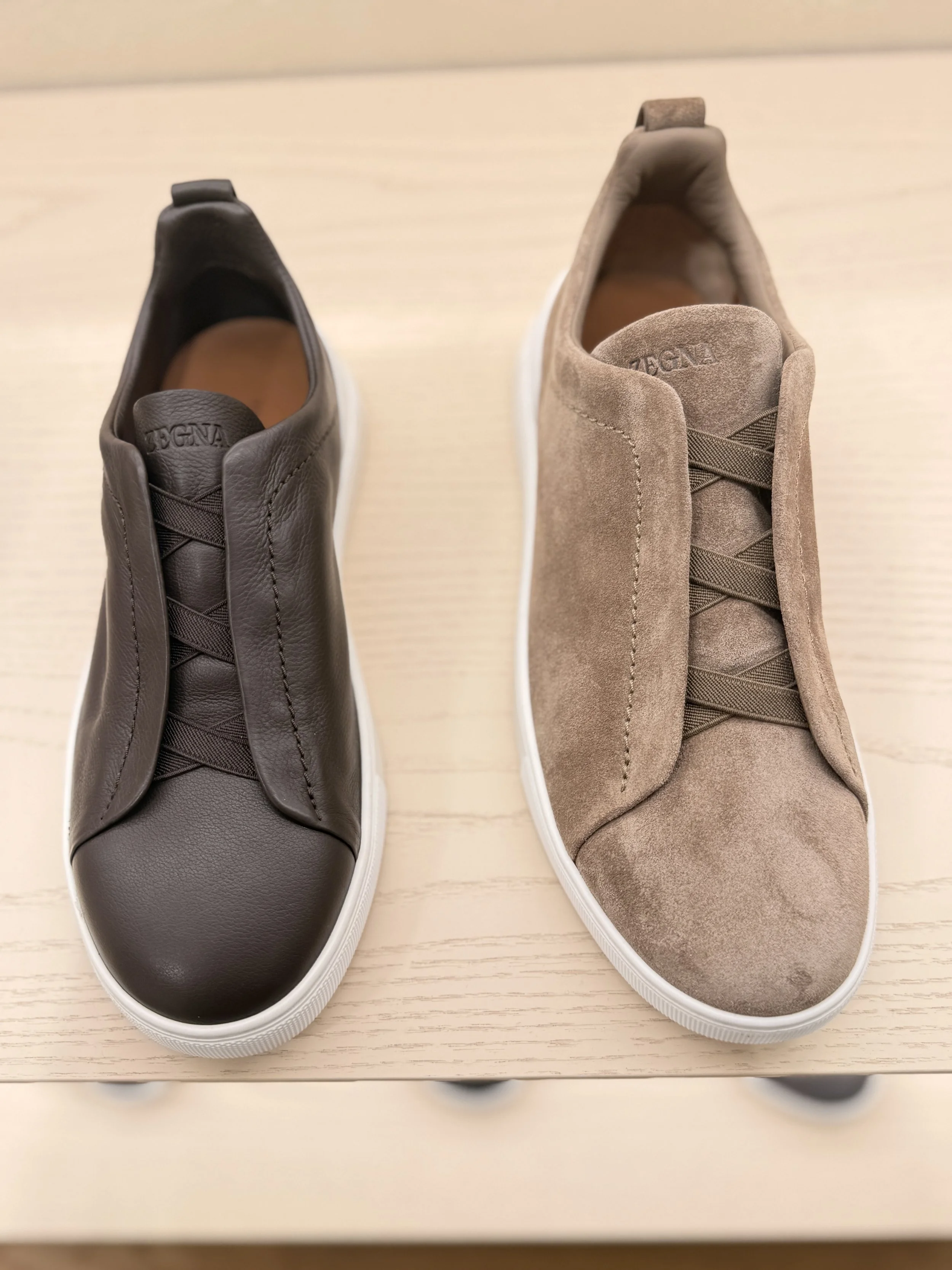Pair of brown leather sneakers on the left and tan suede sneakers on the right, both with white soles, placed on a light wooden surface.