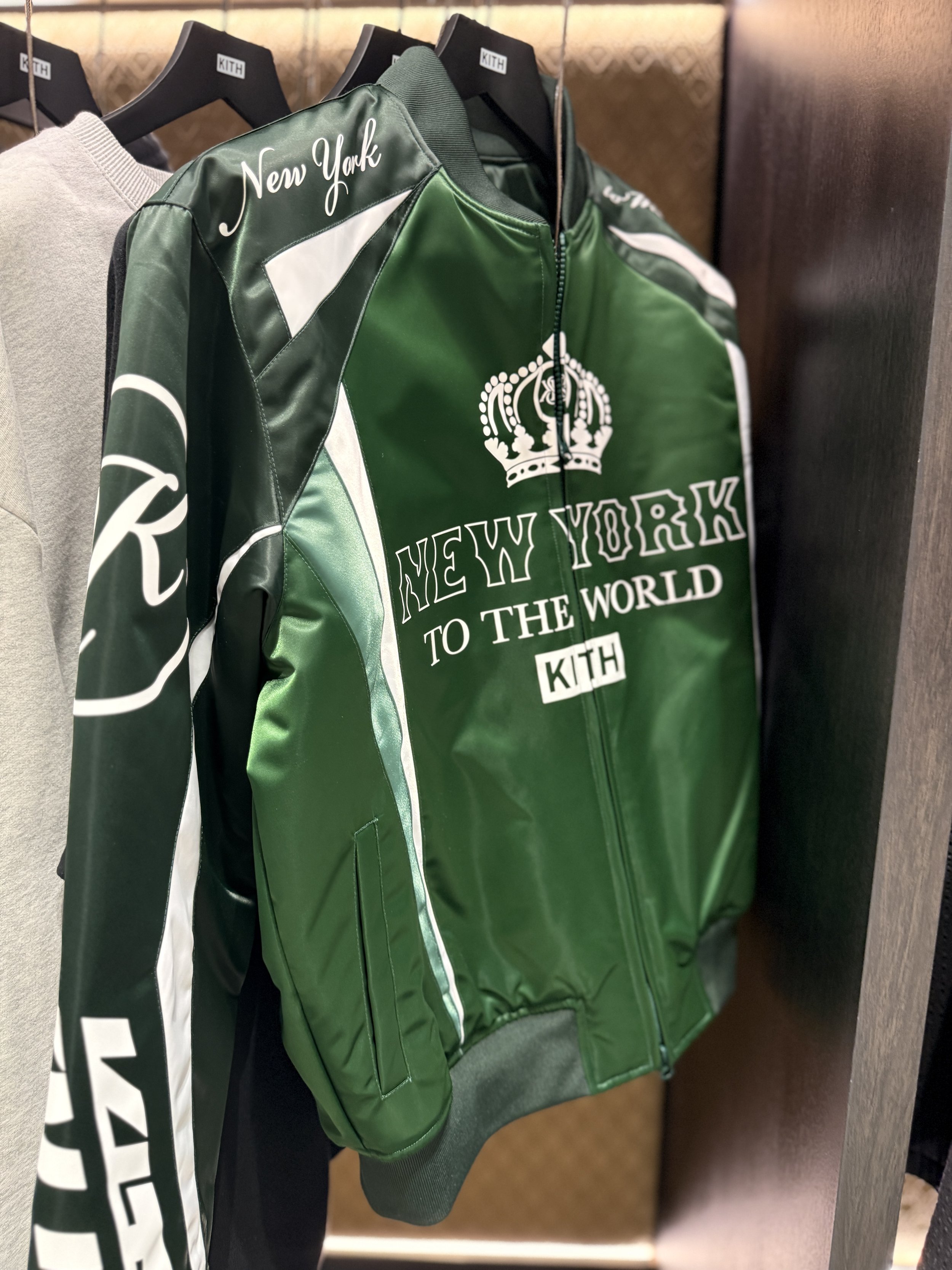 Green Kith brand jacket with white text and a crown graphic, displayed on a hanger in a retail store.