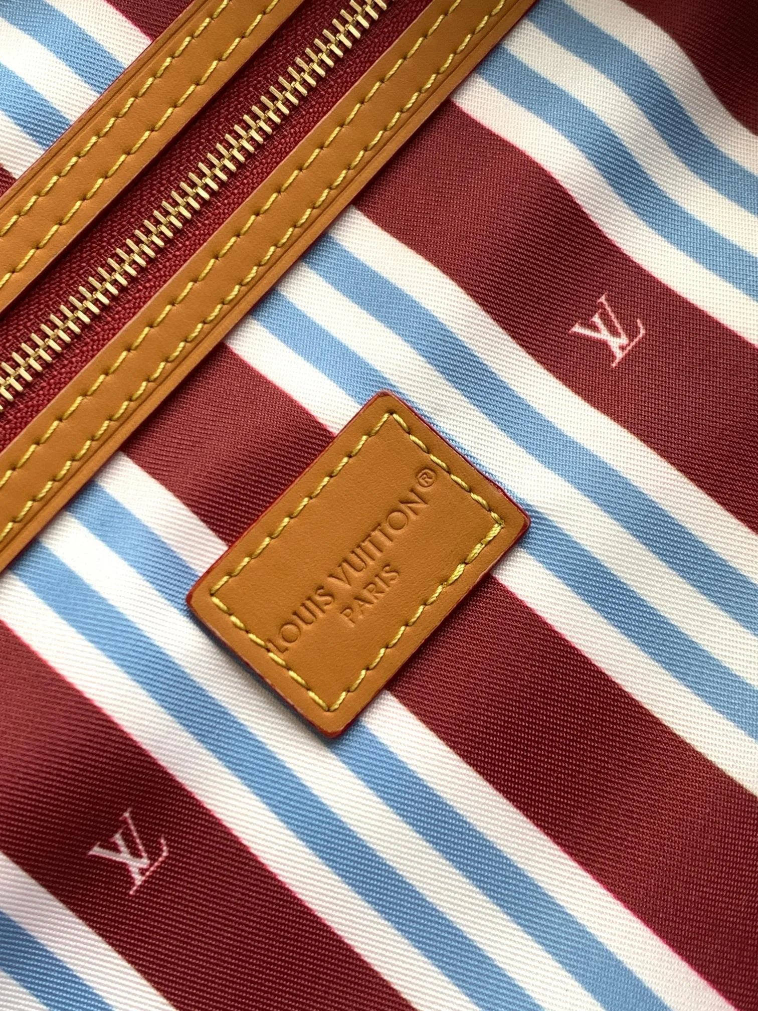 Close-up of a Louis Vuitton bag with red, white, and blue striped fabric, a brown leather patch with yellow stitching that says 'LOUIS VUITTON PARIS', and a gold zipper.