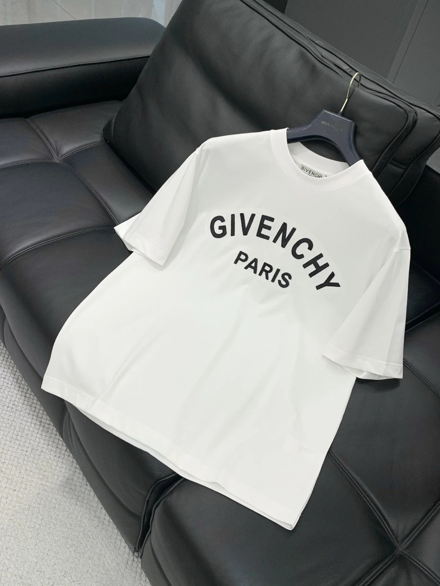 A white Givenchy Paris T-shirt hanging on a black hanger on a black leather couch.