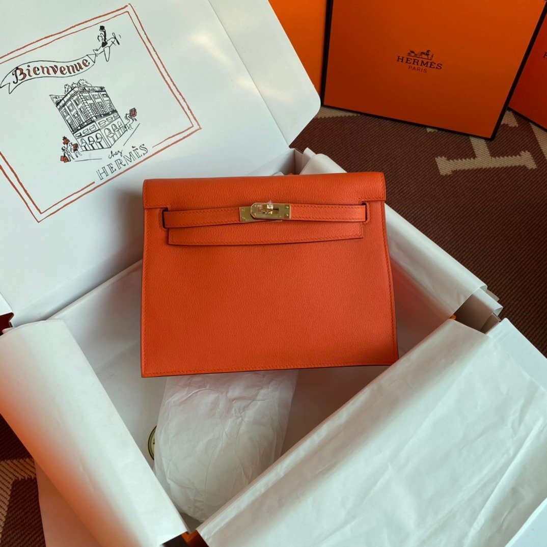 An orange Hermès handbag with a gold clasp inside an open white Hermès box, with Hermès branded packaging and tissue paper nearby.
