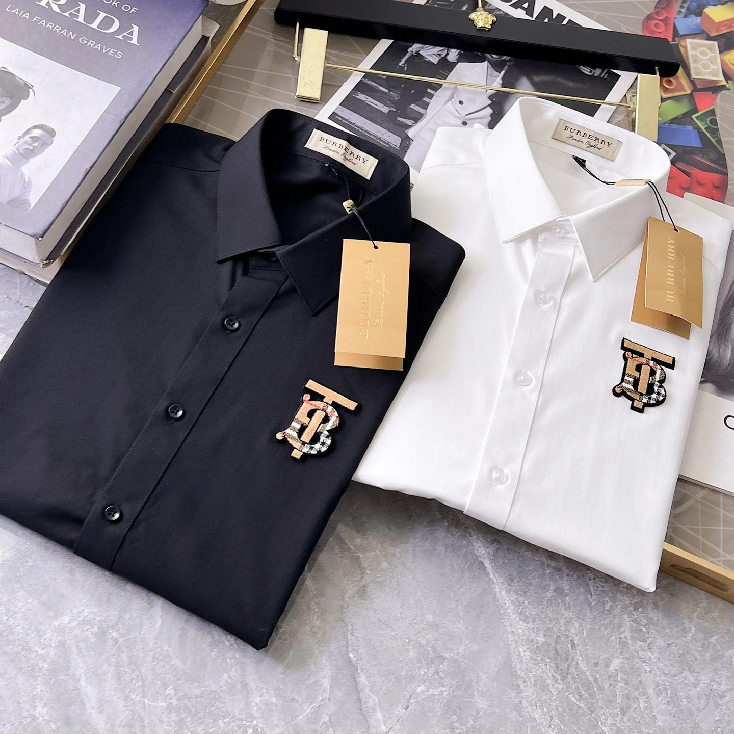 Two neatly folded designer shirts, one black and one white, with Burberry tags and embroidered logos on the chest, are displayed on a surface along with fashion books and accessories.