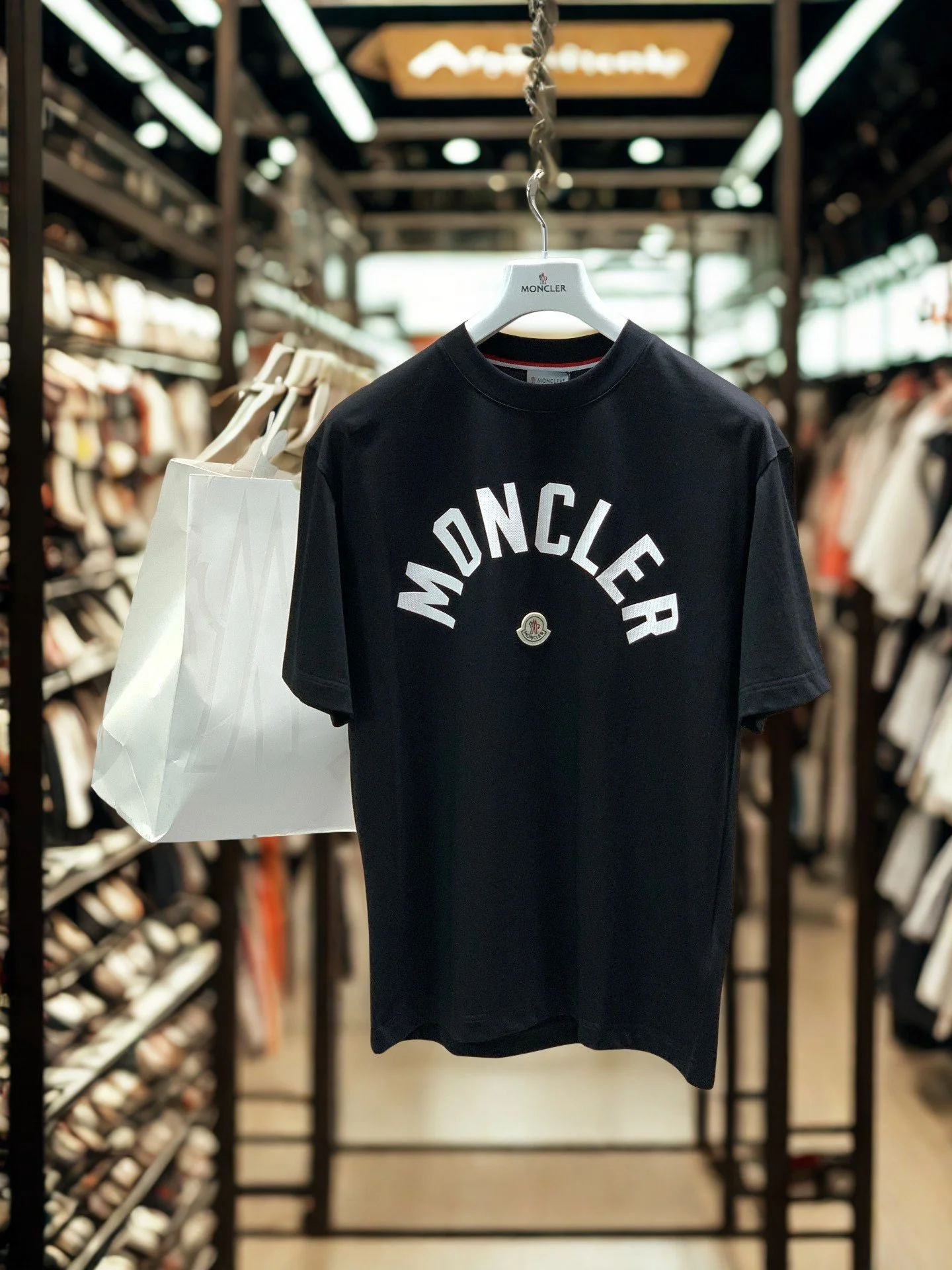 Black Moncler T-shirt with large white lettering hanging on a rack in a store, with shelves of shoes on either side.