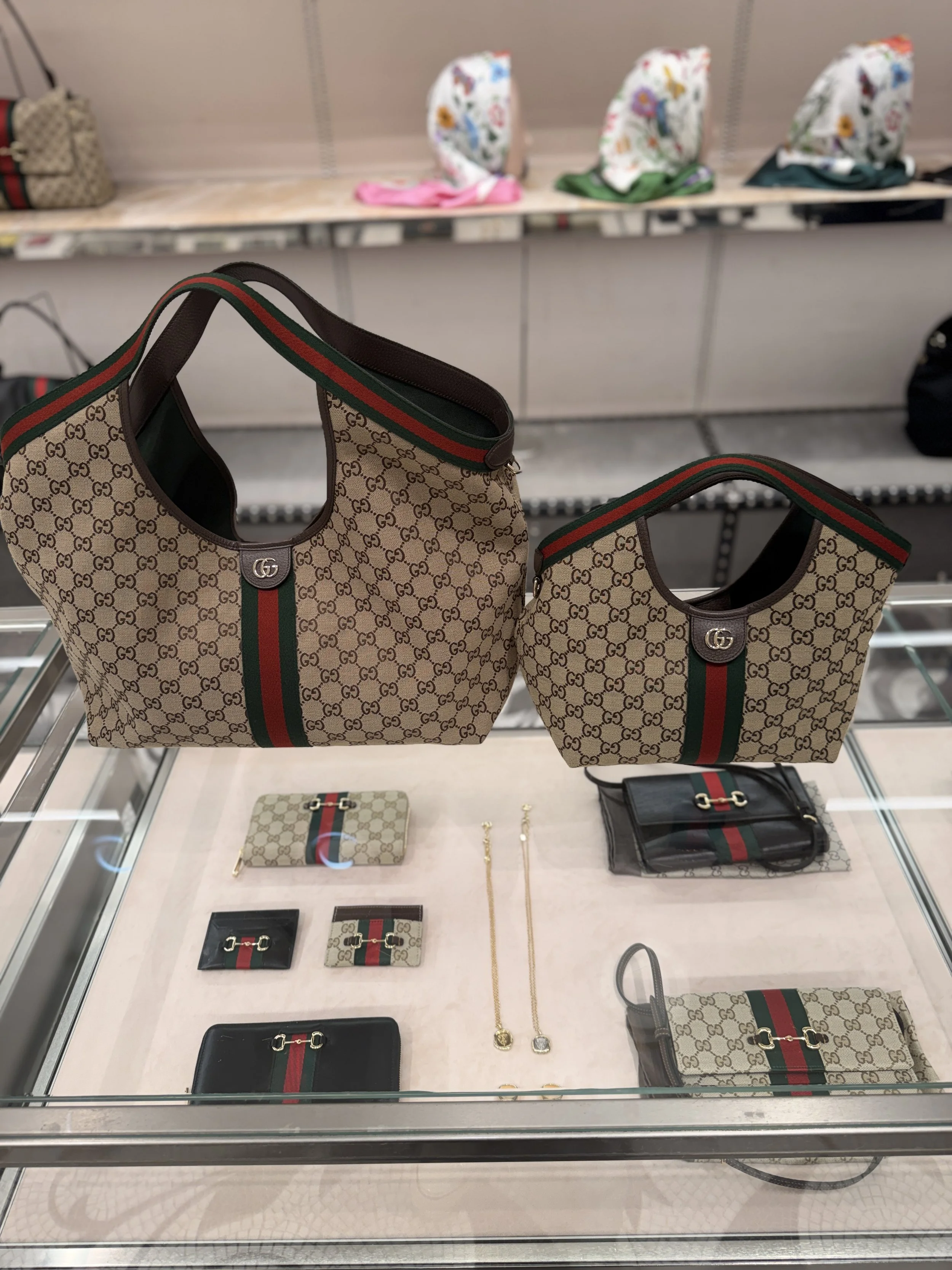 Display of Gucci handbags and accessories featuring the brand's signature monogram pattern, with green and red striped accents, placed in a glass showcase in a retail store.