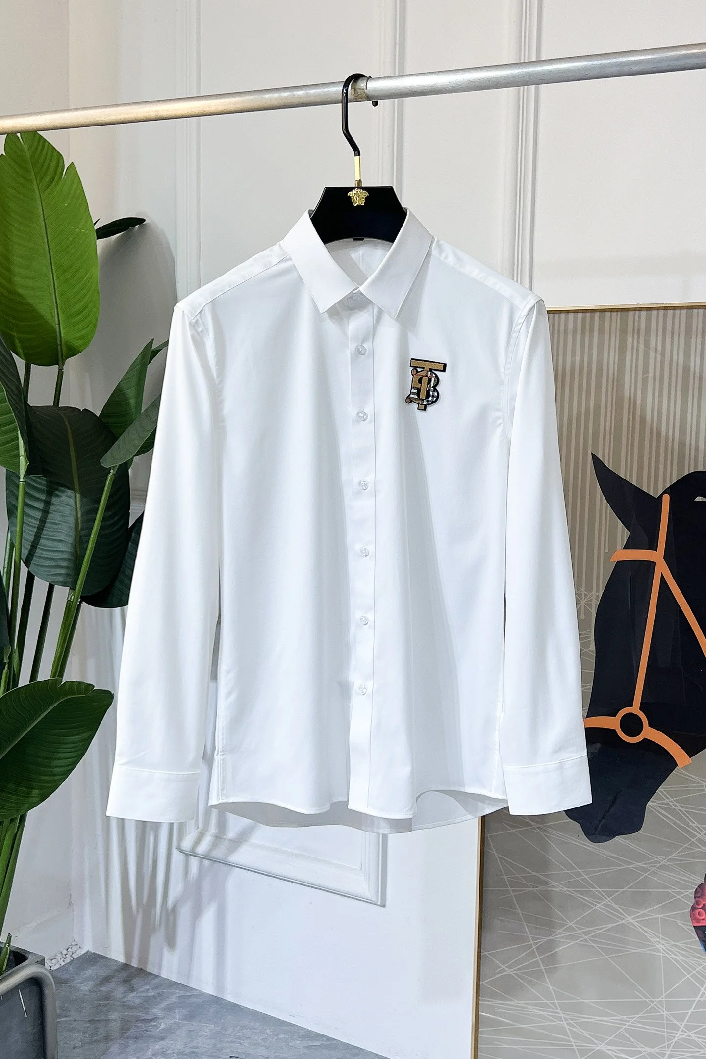 White button-up shirt with a 'TB' logo pin on the left chest, hanging on a black hanger from a metal clothing rod, with a green plant and a black horse head sculpture in the background.