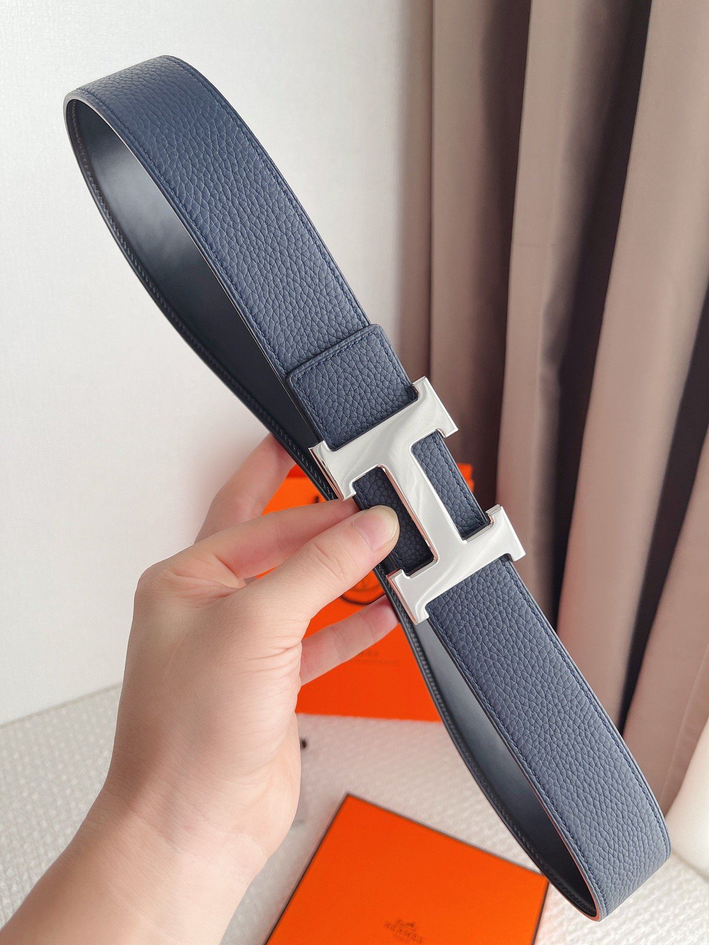 Hand holding a navy blue leather belt with a large silver 'H' buckle.