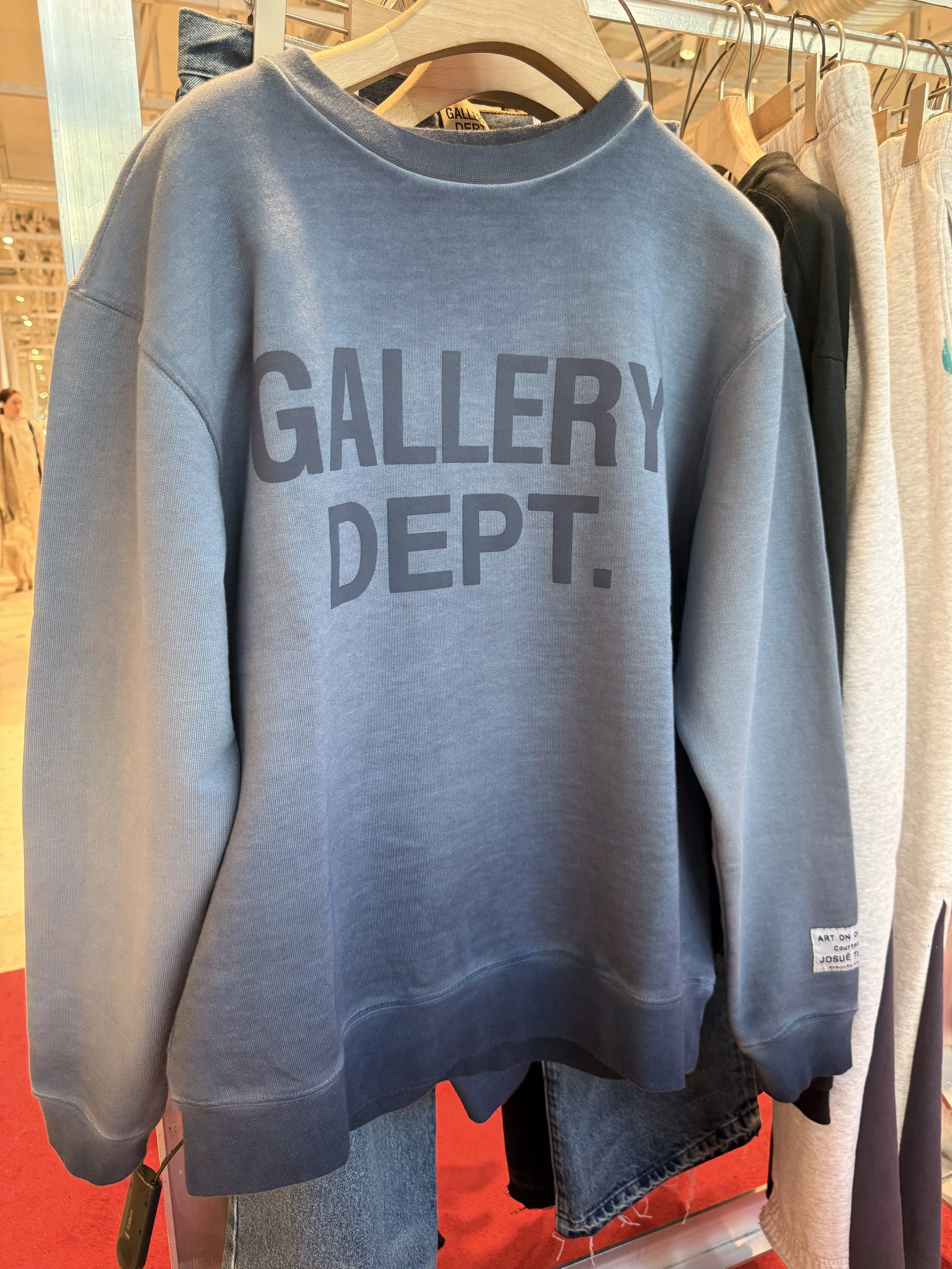 A blue sweatshirt hanging on a wooden hanger in a clothing store. The sweatshirt has the words 'GALLERY DEPT.' printed on the front in dark letters.