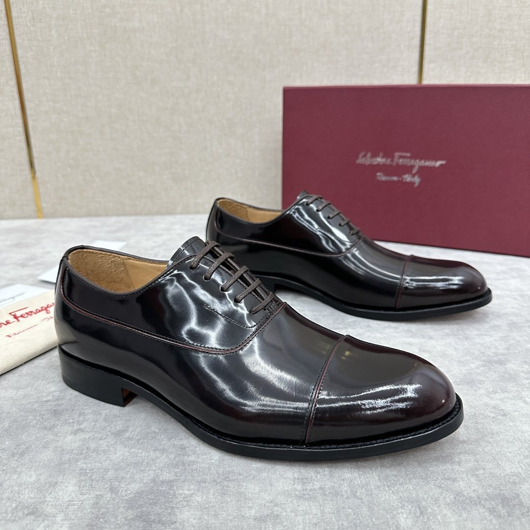 Pair of shiny black leather men's dress shoes on a gray surface with a pink box and a beige cloth bag in the background.