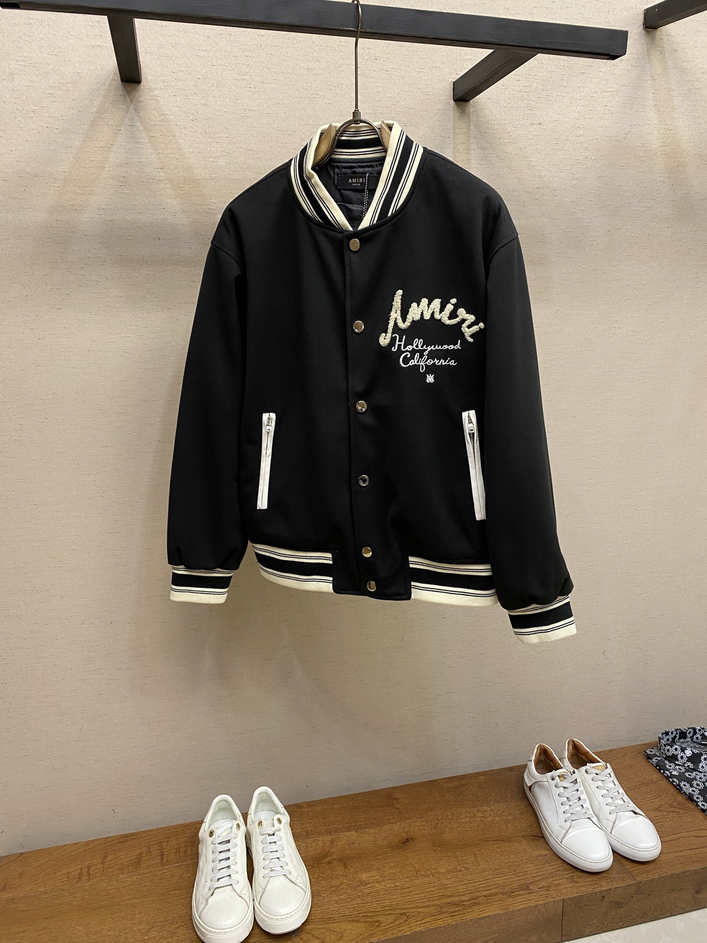 A black varsity jacket with white lettering and striped cuffs, collar, and waistband hanging on a hanger, with white sneakers on a wooden surface below.