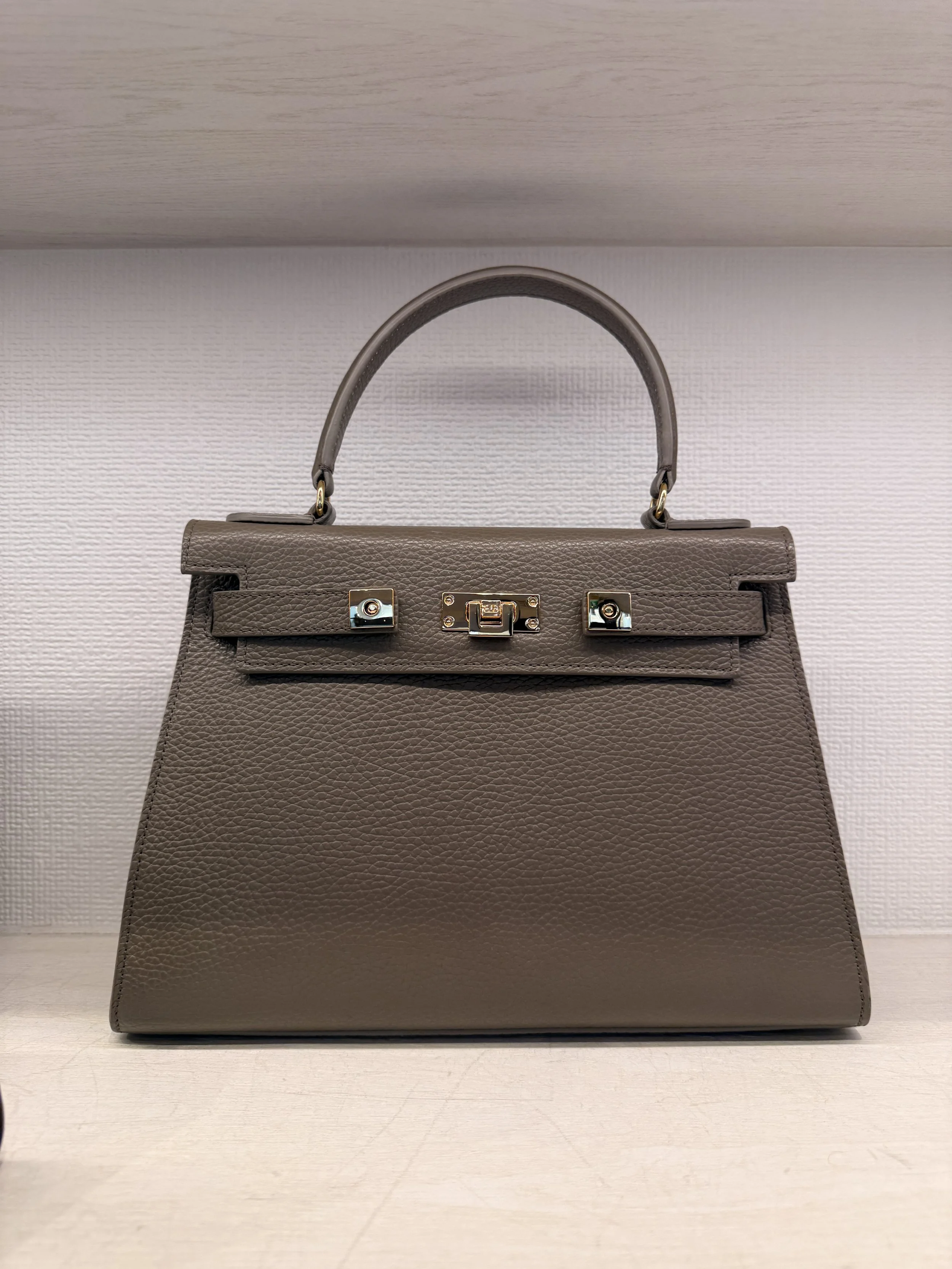 A taupe-colored leather handbag with a top handle, metal hardware closure, and a textured surface, displayed against a white background.