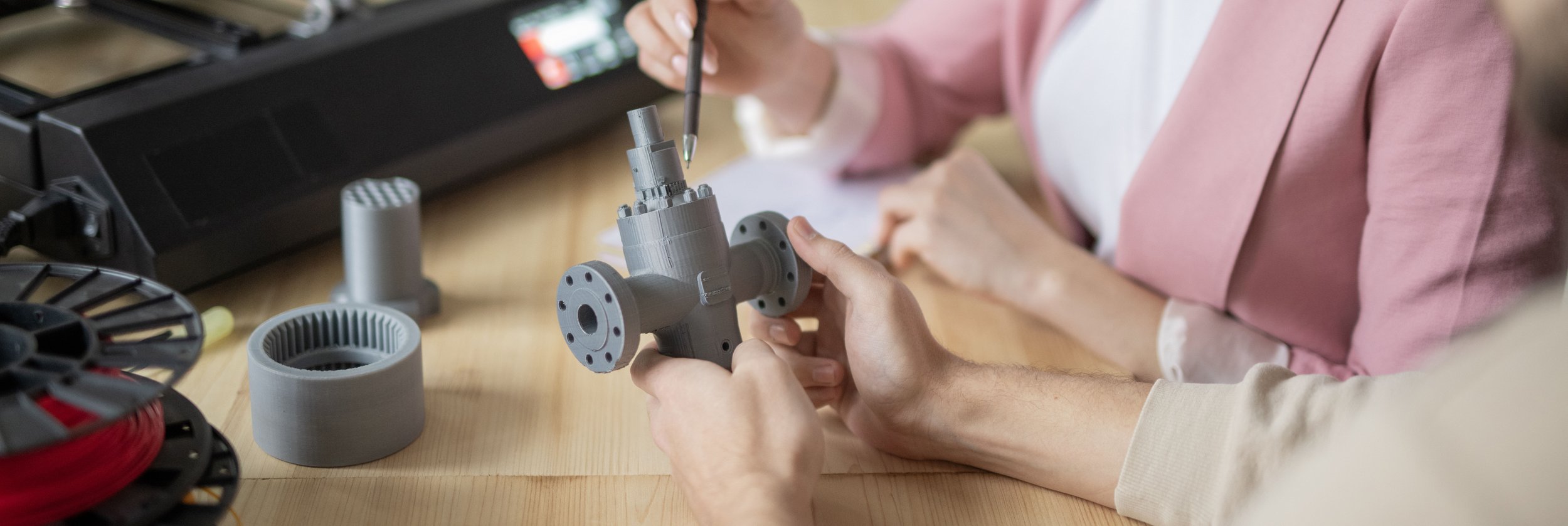 Two people working on a 3D printed object with a gray plastic component, and various tools and 3D printing parts on the wooden table.