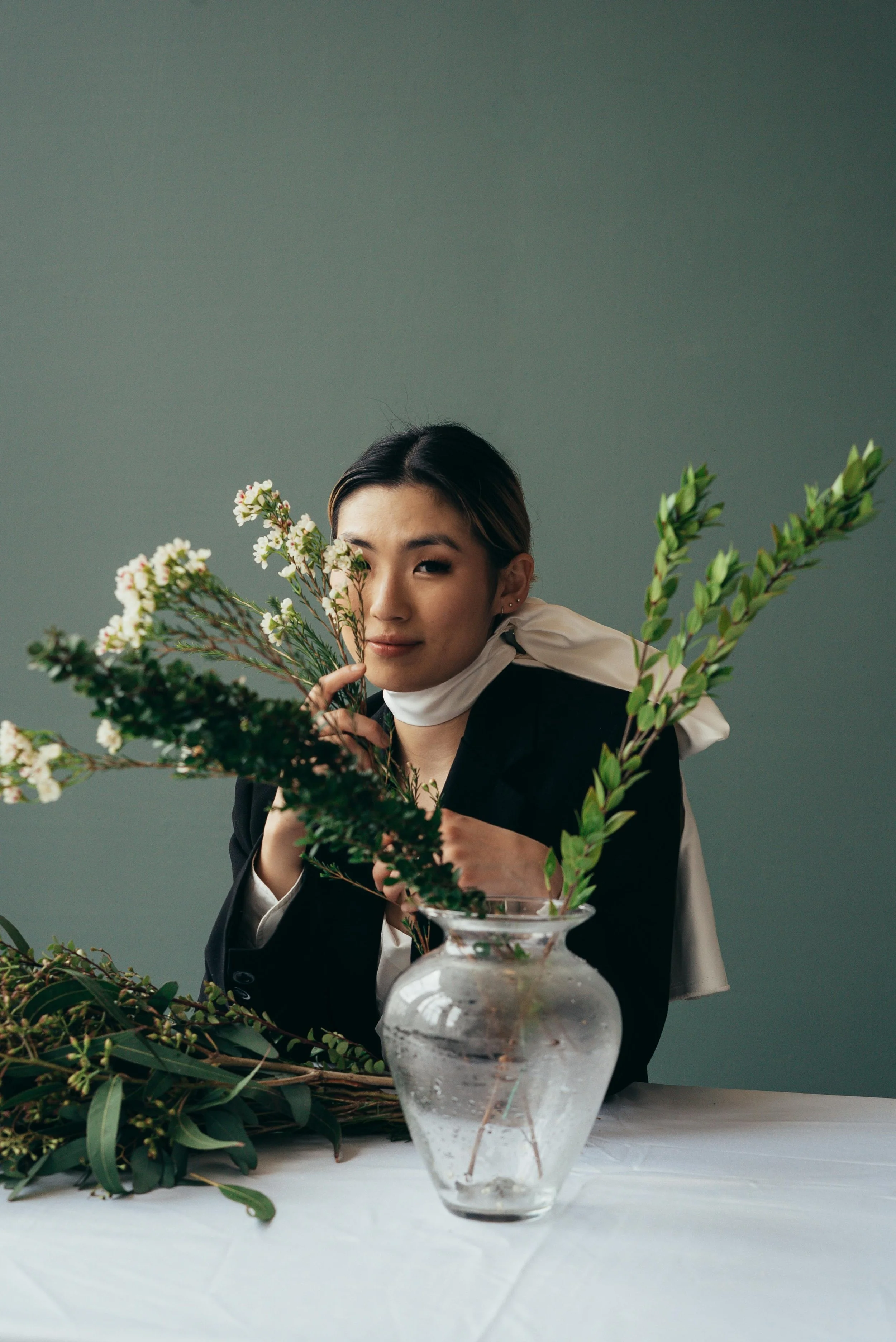 A woman with dark hair, wearing a black jacket and white scarf, stands behind a table with floral arrangements in a clear glass vase. She partly hides her face with a branch of white flowers, smiling and looking at the camera against a plain green background.