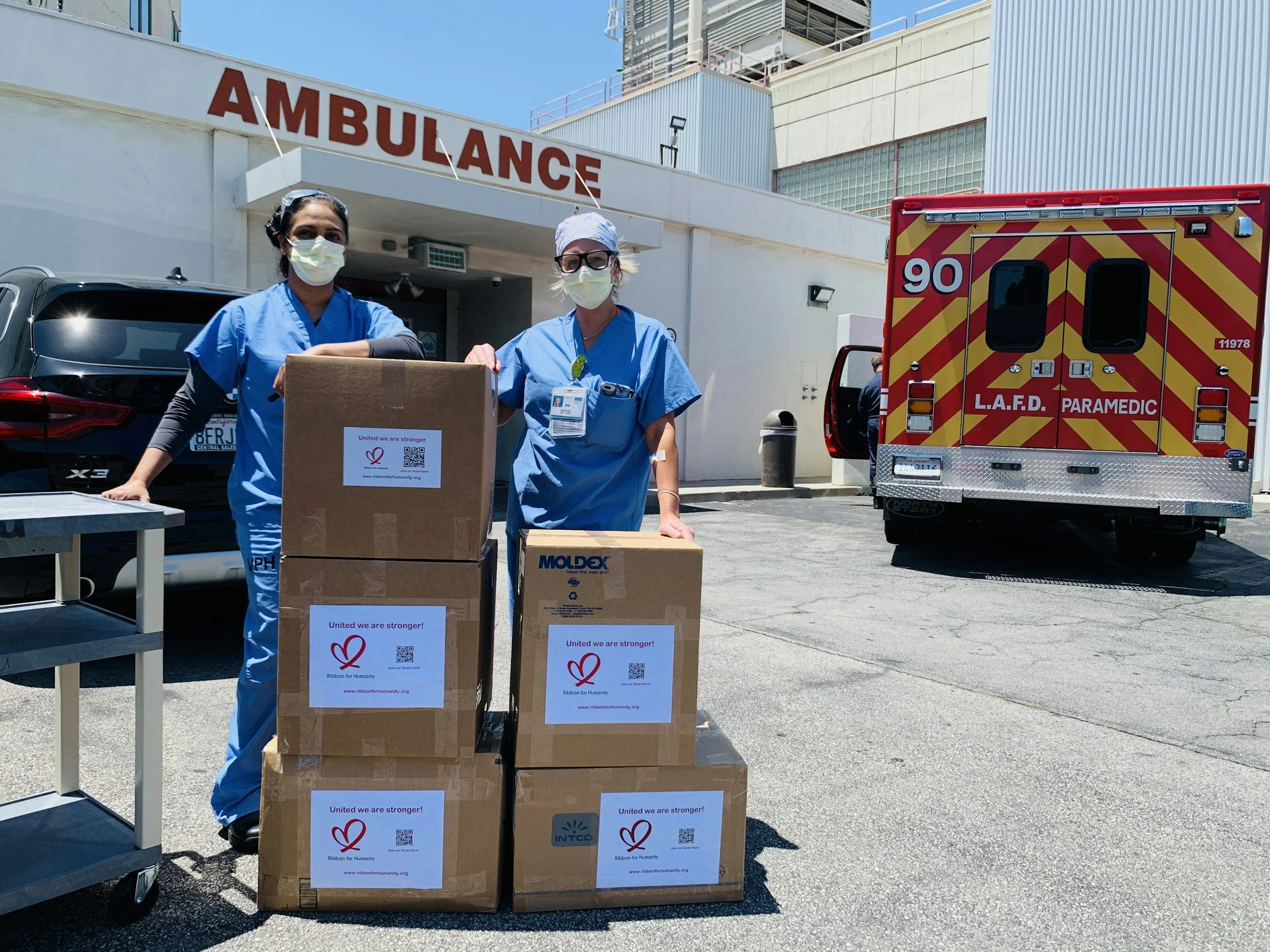 Two healthcare workers in blue scrubs and masks unloading boxes labeled "United we are stronger!" outside a hospital with an ambulance in the background.