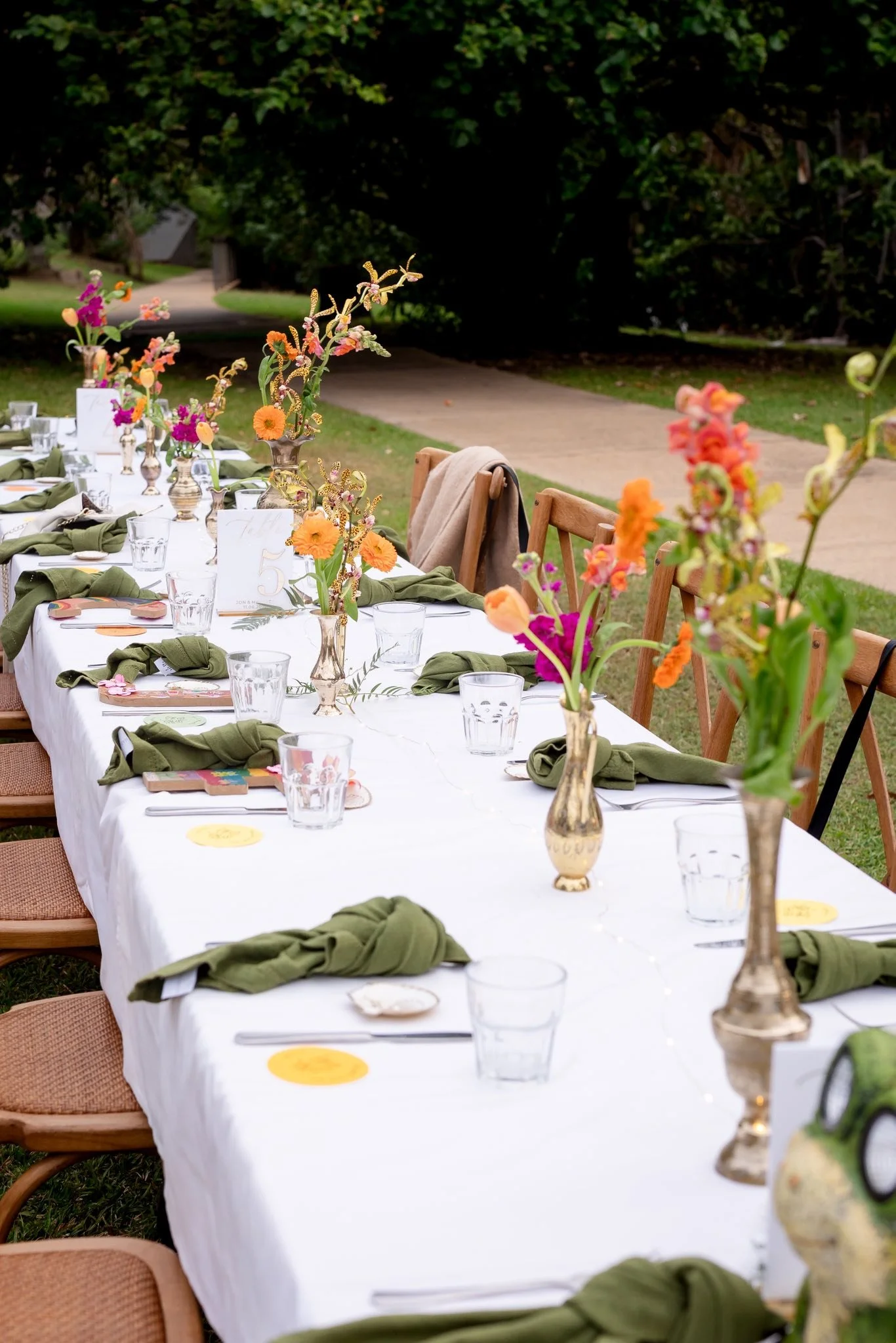 A long outdoor dining table with white tablecloth, green napkins, glasses, and floral centerpieces in gold and glass vases, set in a garden with chairs and a pathway in the background.